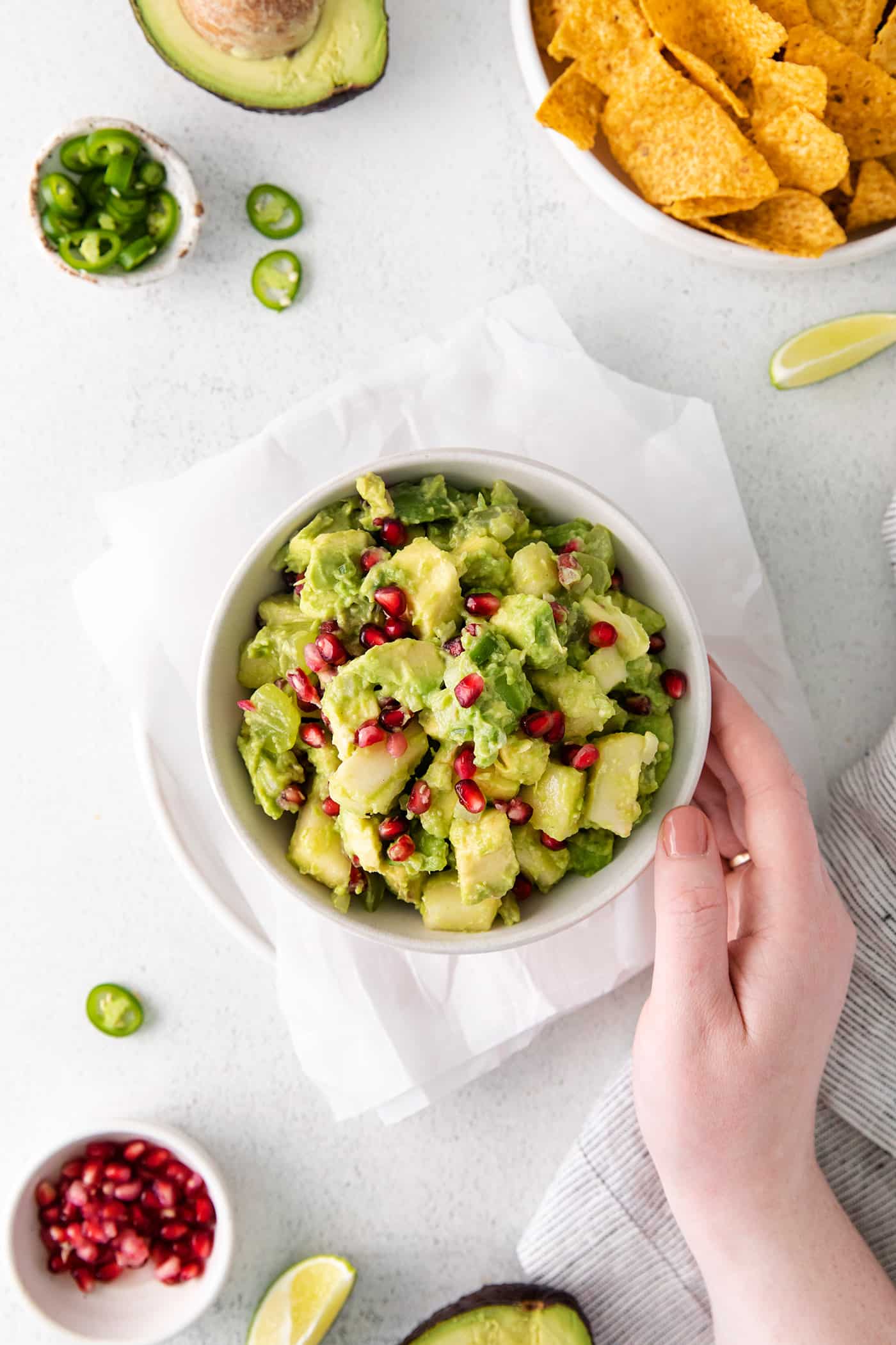 With sweet pops of fruit and pomegranate seeds, this Holiday Guacamole is a quick and festive party appetizer! Delicious with tortilla chips! Overhead view of a hand touching a white bowl of holiday guacamole, next to a bowl of tortilla chips.