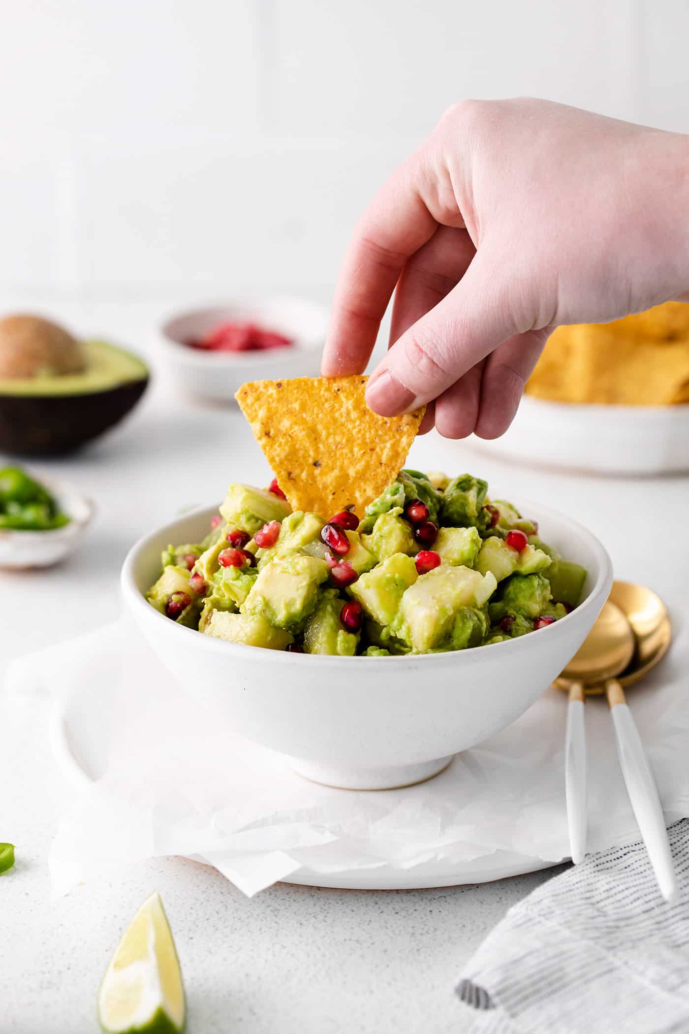 With sweet pops of fruit and pomegranate seeds, this Holiday Guacamole is a quick and festive party appetizer! Delicious with tortilla chips! A hand dipping a chip into a bowl of holiday guacamole.
