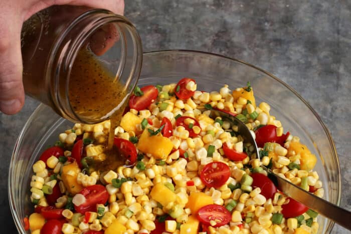 With fresh corn, mangos, tomatoes, and a touch of heat, plus a light and flavorful dressing, this Sweet Corn Salad is a summer treat! pouring dressing over the fresh ingredients in a serving bowl