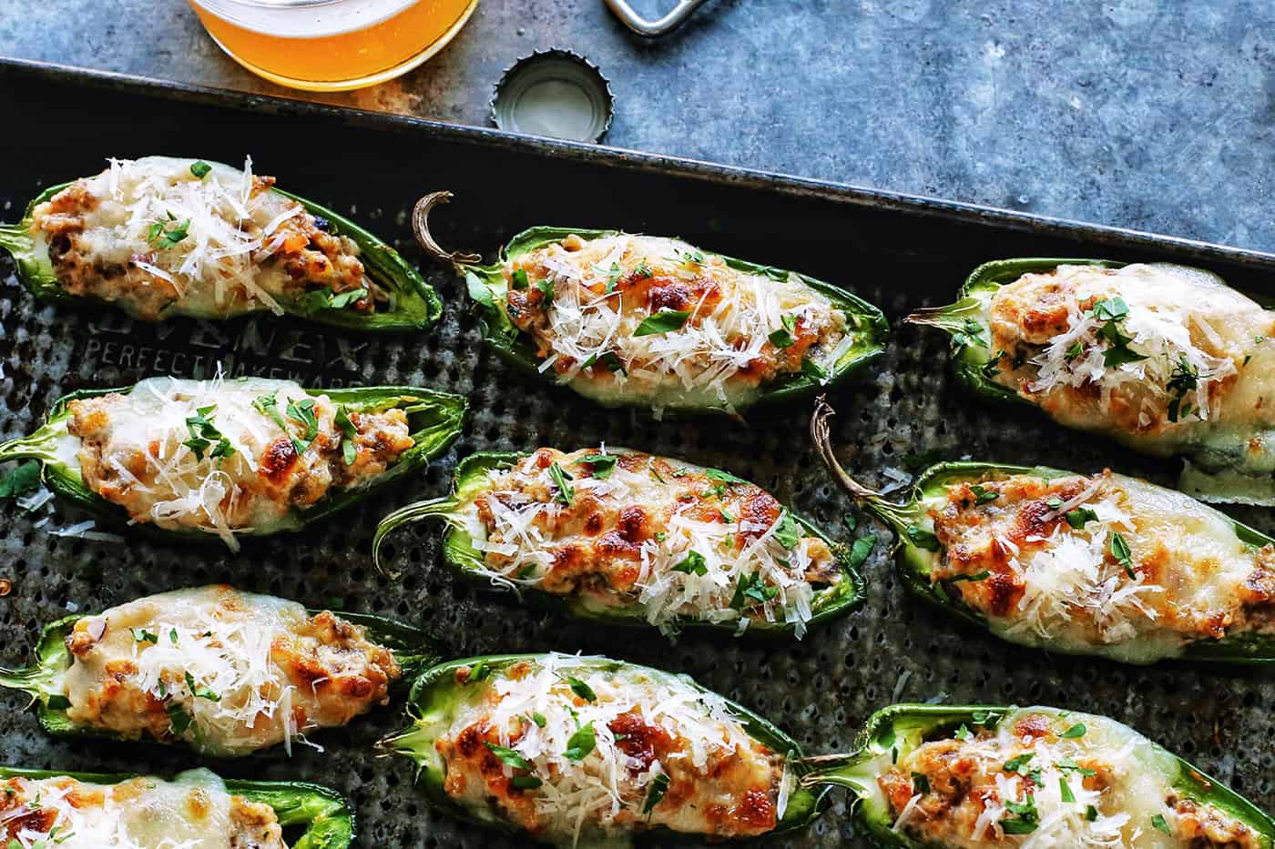 With Italian sausage, cream cheese, Parmesan, and mozzarella, these easy stuffed jalapeños are extra cheesy with the perfect amount of heat! stuffed jalapeno peppers on a baking pan, plus a glass of beer