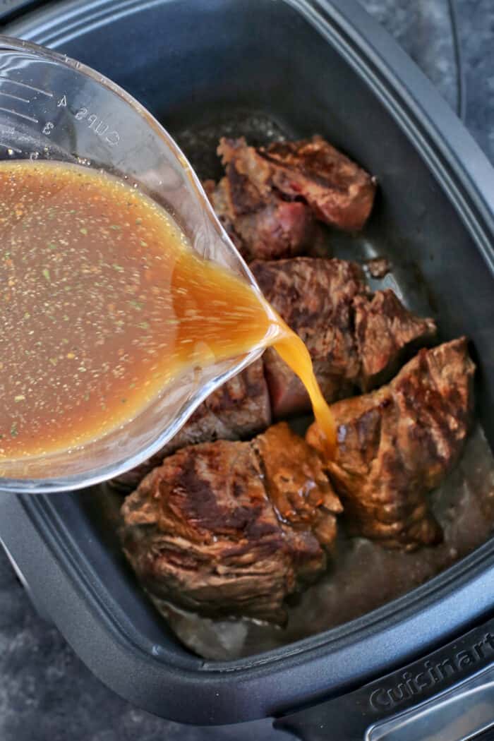 Italian seasoning and beef broth being poured over chuck roast inside the slow cooker