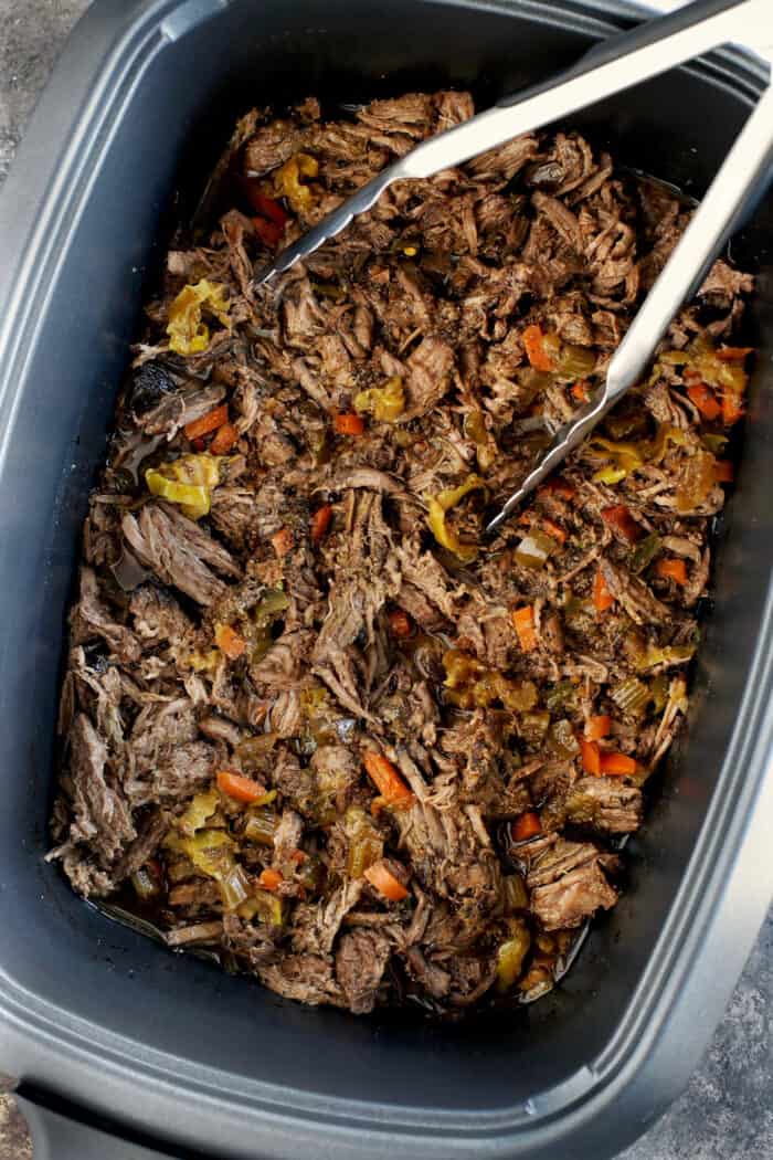 Tongs resting in shredded Italian beef inside the slow cooker