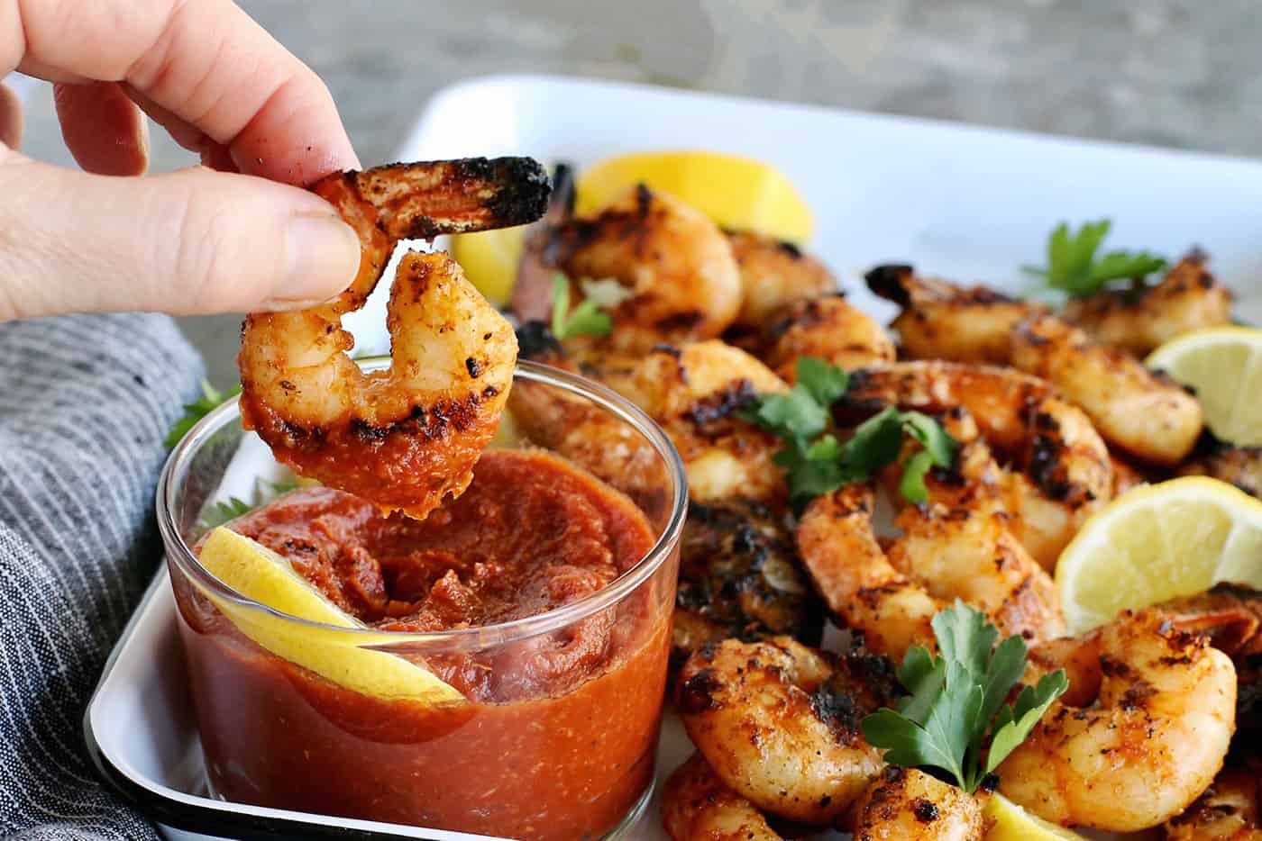 Tossed with olive oil and a few spices, this easy shrimp cocktail is perfectly flavored and grilled until just charred. So good! A hand dipping a grilled shrimp in cocktail sauce.