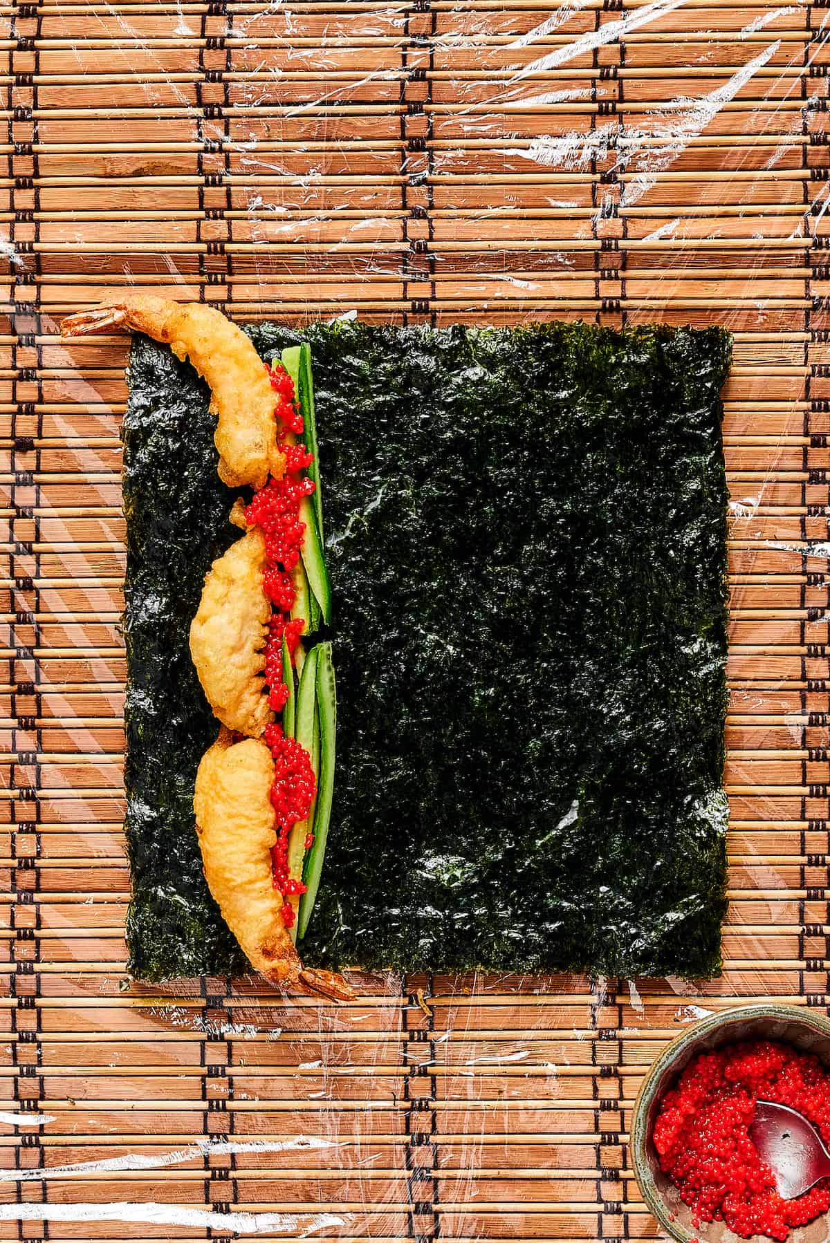 This dragon roll features tempura shrimp, cucumber, tobiko, and avocado wrapped in nori and rice. It's easy to make and such a crowd-pleaser! Cucumber, shrimp, and tobiko on a sheet of nori.