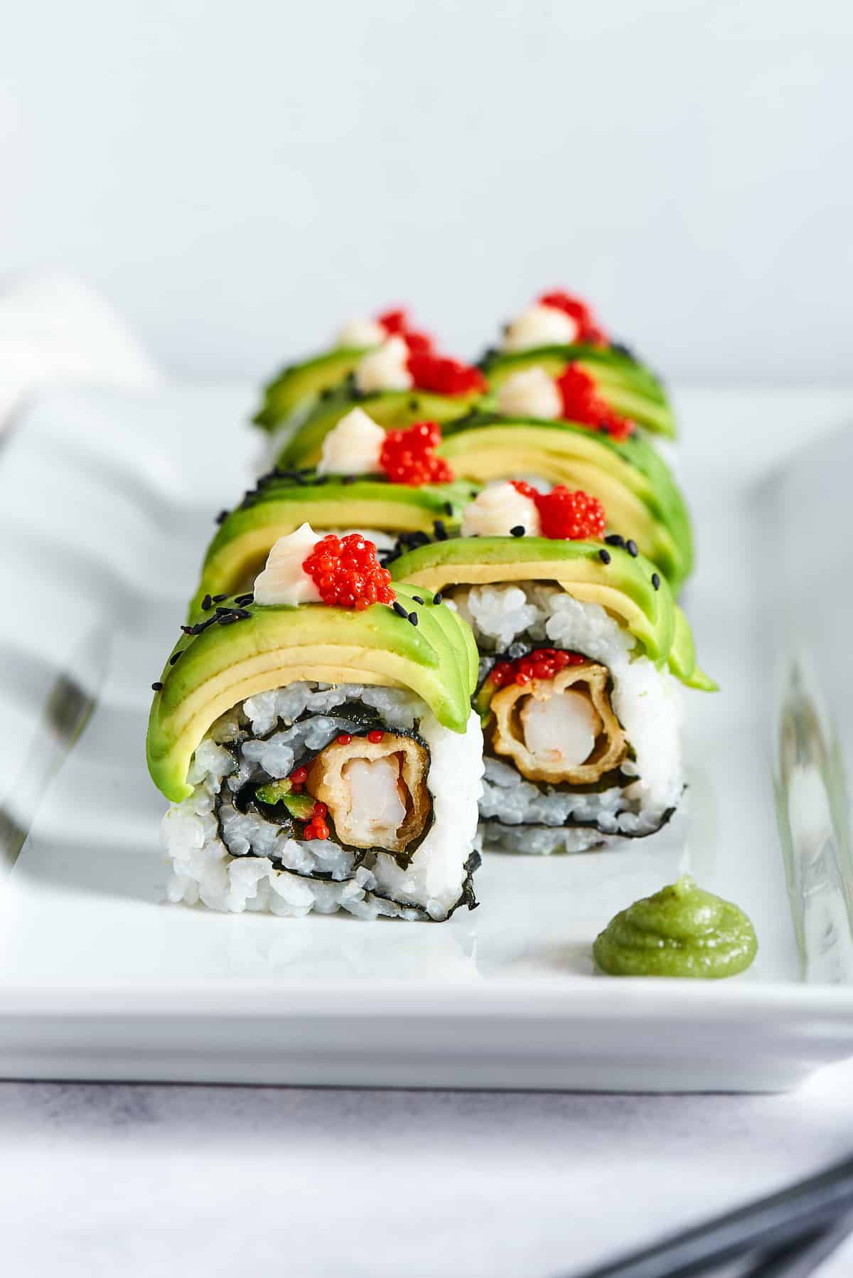 This dragon roll features tempura shrimp, cucumber, tobiko, and avocado wrapped in nori and rice. It's easy to make and such a crowd-pleaser! Front view of a dragon roll on a white plate.