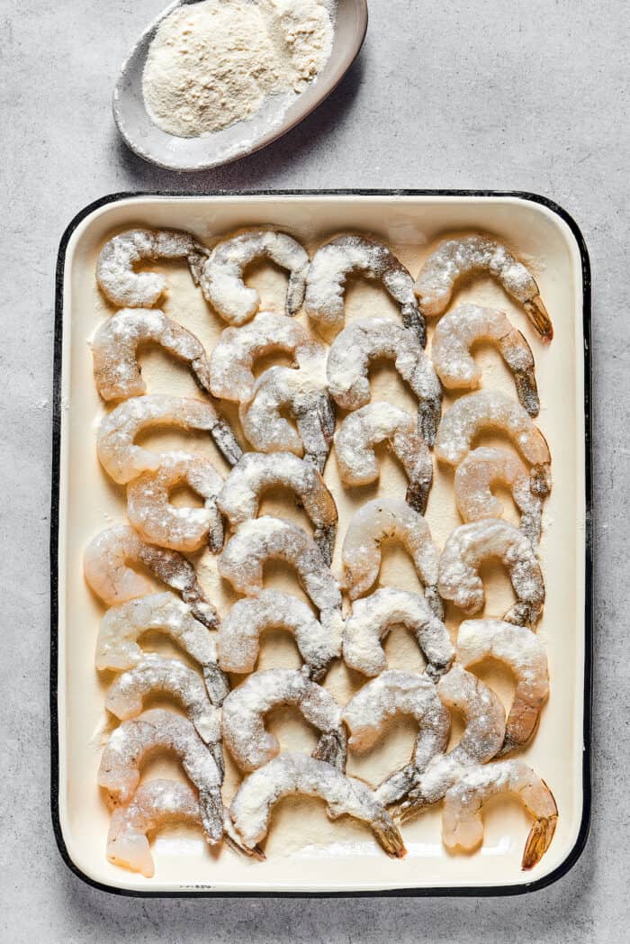 This easy shrimp tempura recipe features juicy, tender shrimp dipped in a light tempura batter. It's then deep-fried until perfectly crispy! Shrimp coated in flour on a baking sheet
