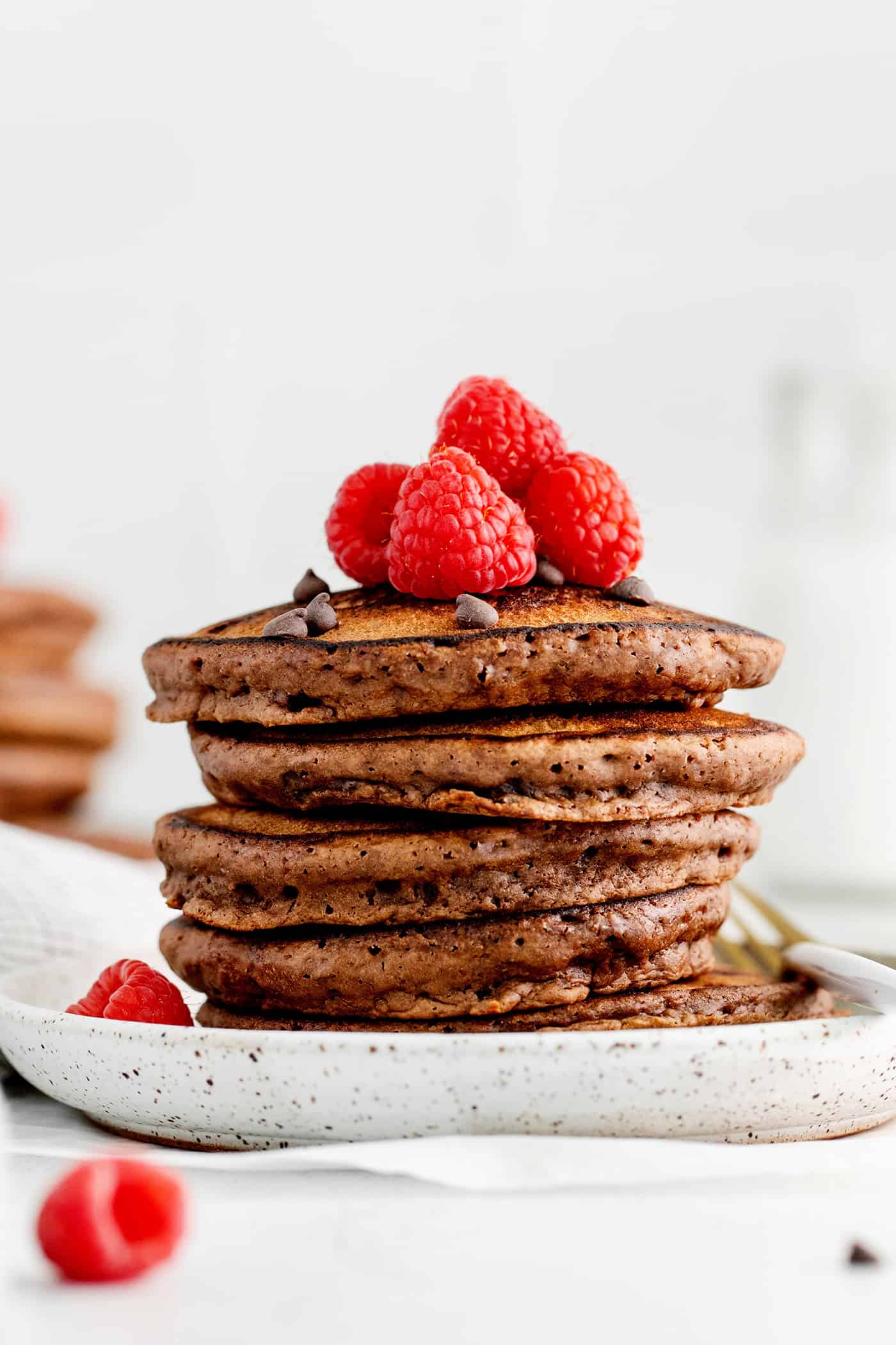Fluffy, fudgy chocolate pancakes are rich in flavor with pockets of melty chocolate chips throughout. The real chocolate lover's breakfast! A stack of chocolate pancakes topped with raspberries on a white plate