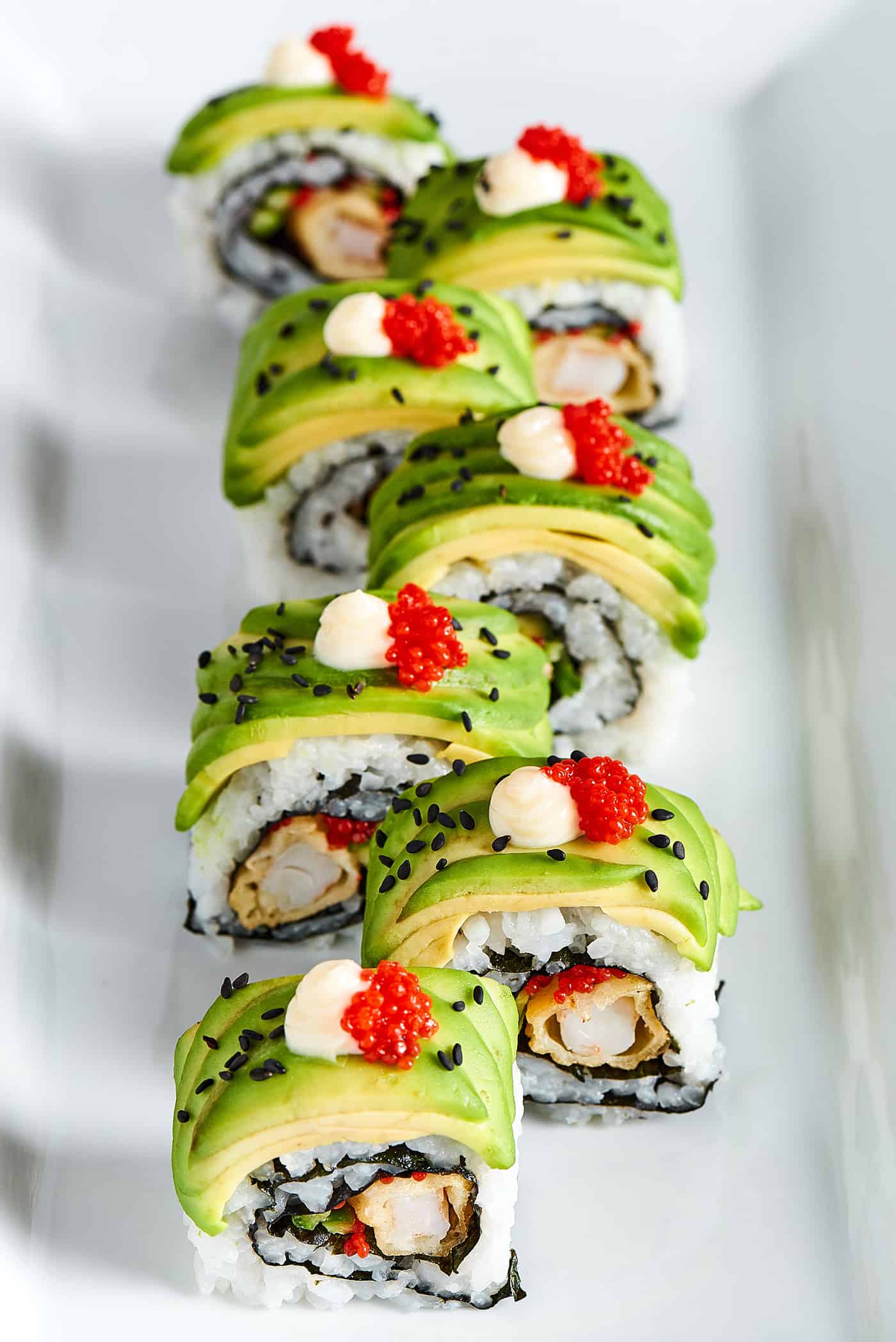 This dragon roll features tempura shrimp, cucumber, tobiko, and avocado wrapped in nori and rice. It's easy to make and such a crowd-pleaser! Overhead photo of sushi roll topped with thin slices of avocado.
