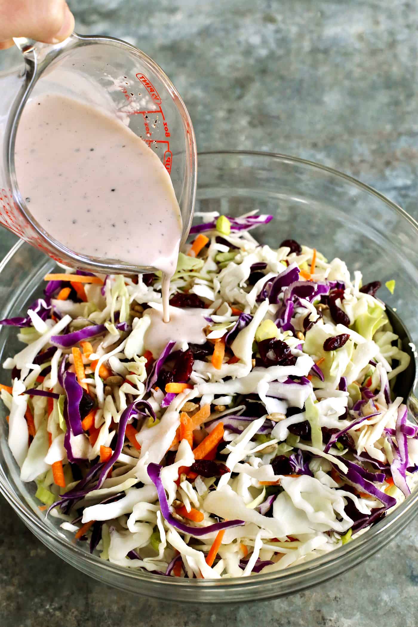 Made with a creamy, tangy dressing, dried cranberries, and sunflower kernels, this Cranberry Coleslaw is an easy side for any occasion. Dressing being poured over a bowl of coleslaw