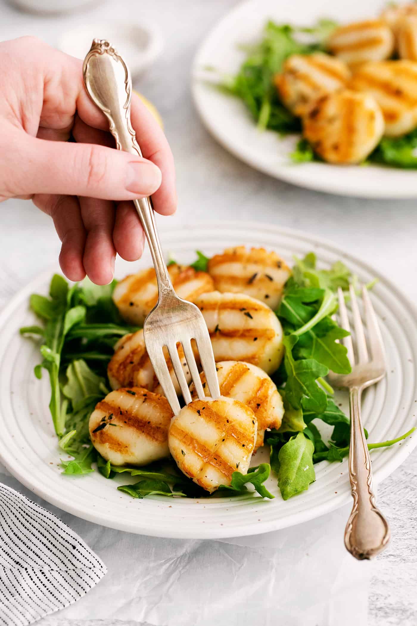 These easy grilled sea scallops are done in 20 minutes and richly flavored with garlic butter and lemon. Cook on an indoor or outdoor grill! A fork in a grilled scallop over a bed of greens