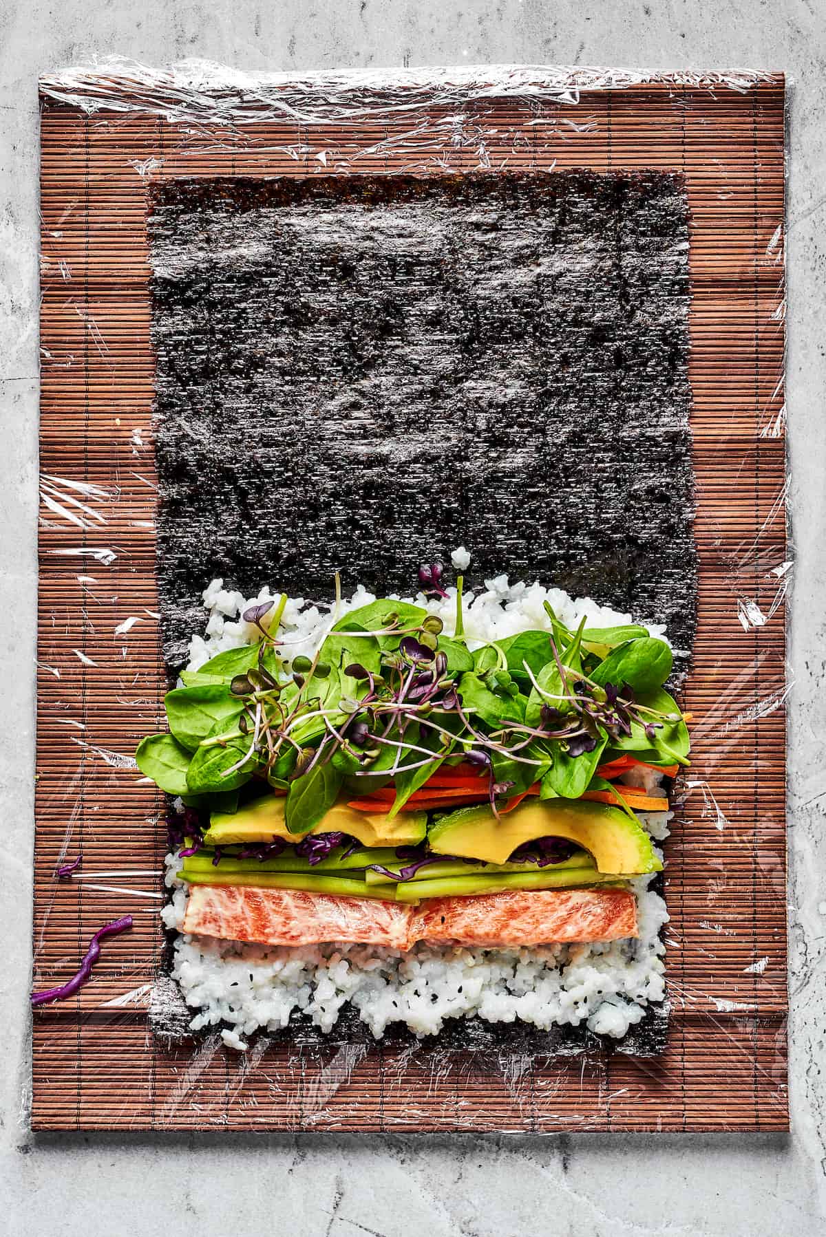 This Sushi Burrito recipe results in a giant sushi roll stuffed with spicy salmon, rice, avocado, carrots, and other veggies. Overhead view of a salmon, rice, and veggies on nori sheets