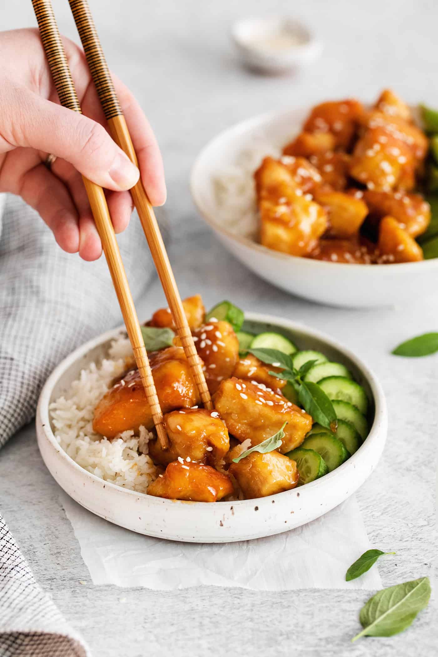 These Honey Garlic Chicken Bowls are quick and easy with moist chunks of chicken tossed in a sweet and sticky garlic sauce. Chopsticks grabbing a piece of honey garlic chicken from a rice bowl