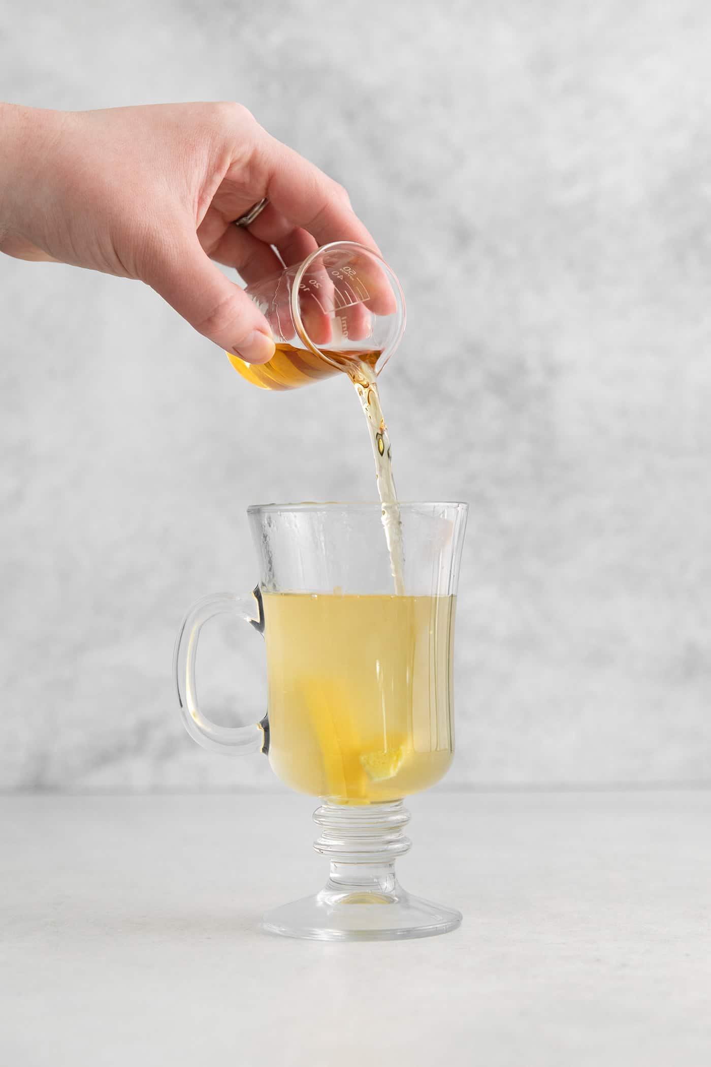 This Hot Toddy is the perfect winter drink! It’s warm and cozy with a hint of earthy honey, spicy ginger, and a burst of lemon. Bourbon being poured into a glass mug with lemon honey water