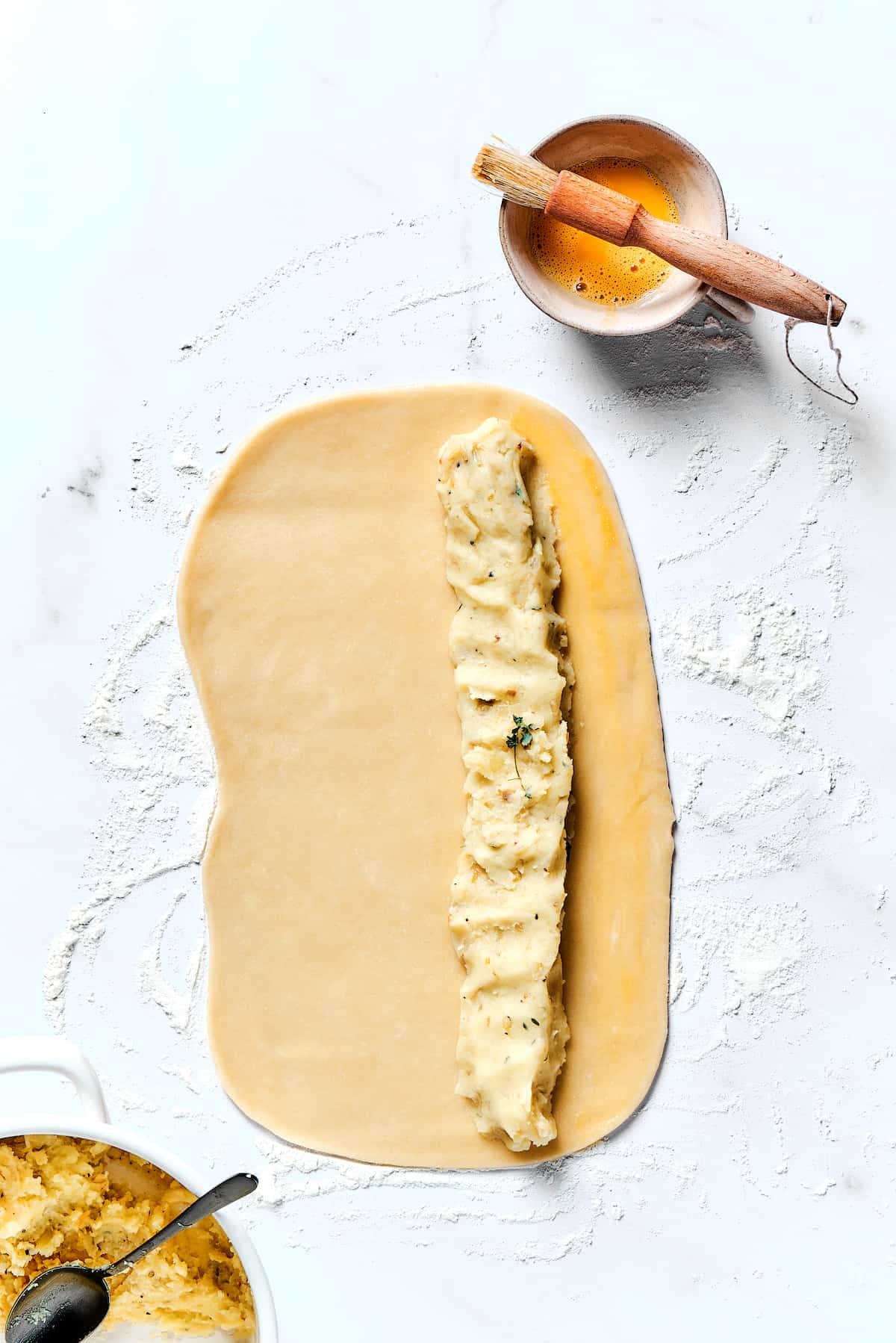 Potato Knishes are a popular Jewish recipe featuring a mashed potato and onion filling wrapped in a thin, flaky dough and baked to golden. A rectangle of dough with a row of mashed potato mixture on part of it