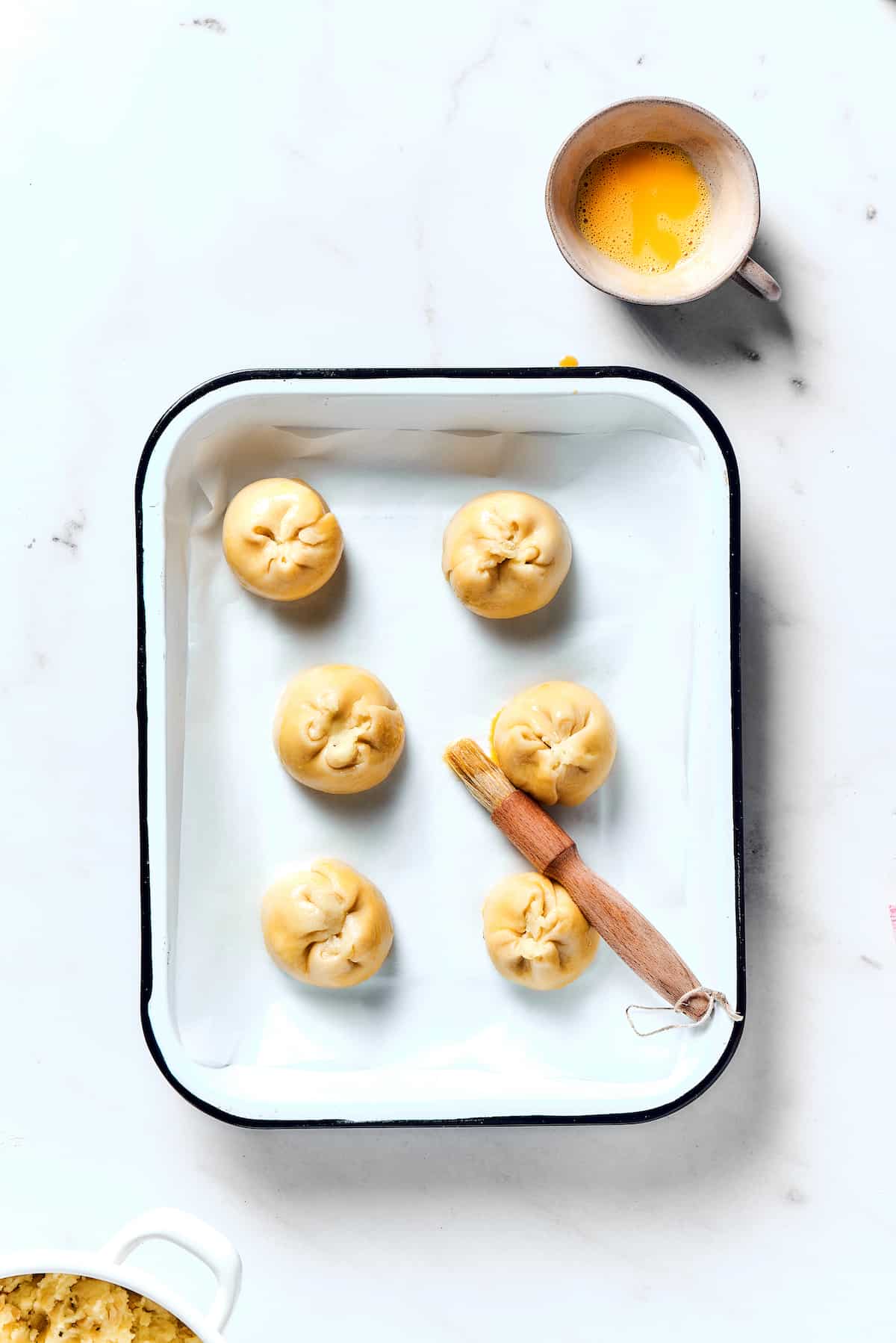 Potato Knishes are a popular Jewish recipe featuring a mashed potato and onion filling wrapped in a thin, flaky dough and baked to golden. Butter being brushed on uncooked knish