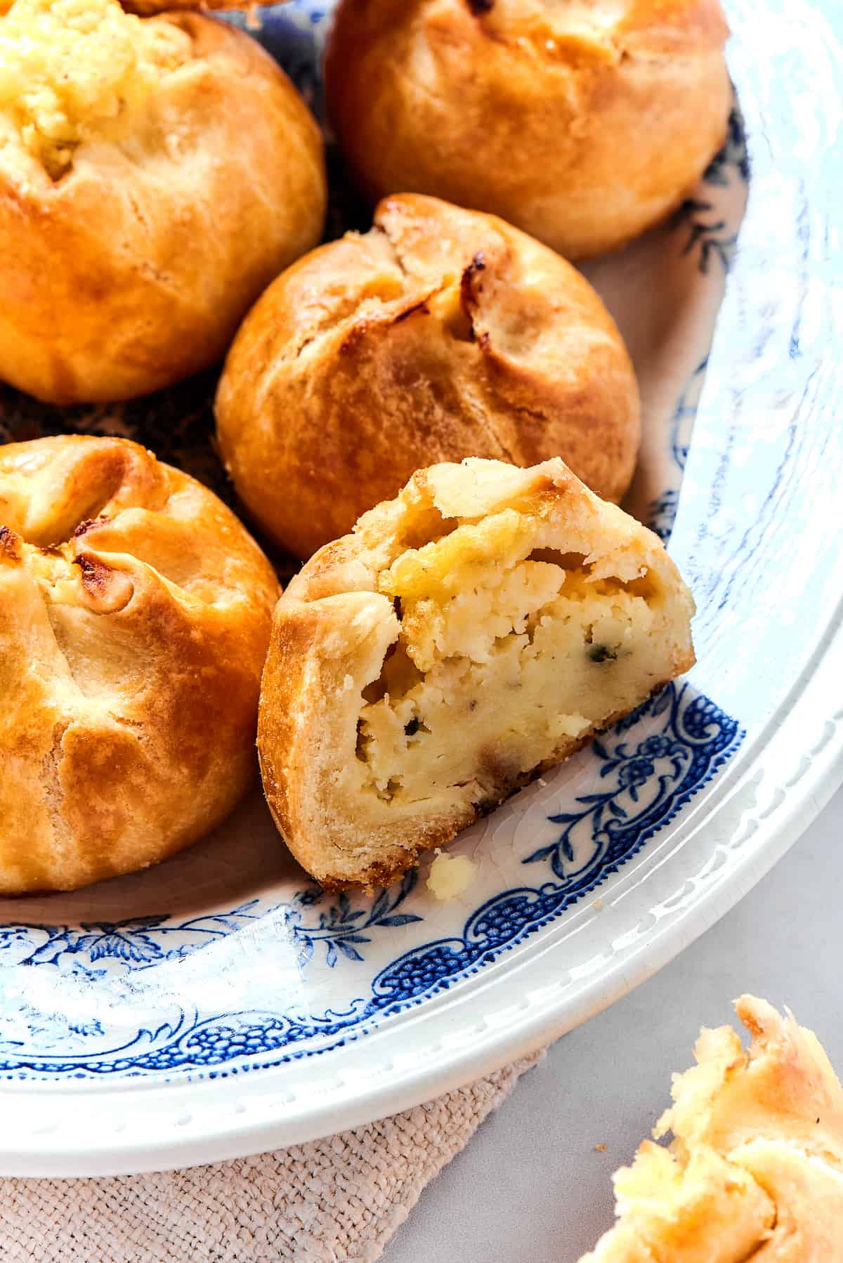 Potato Knishes are a popular Jewish recipe featuring a mashed potato and onion filling wrapped in a thin, flaky dough and baked to golden. A corner of a plate of knish, with one cut in half