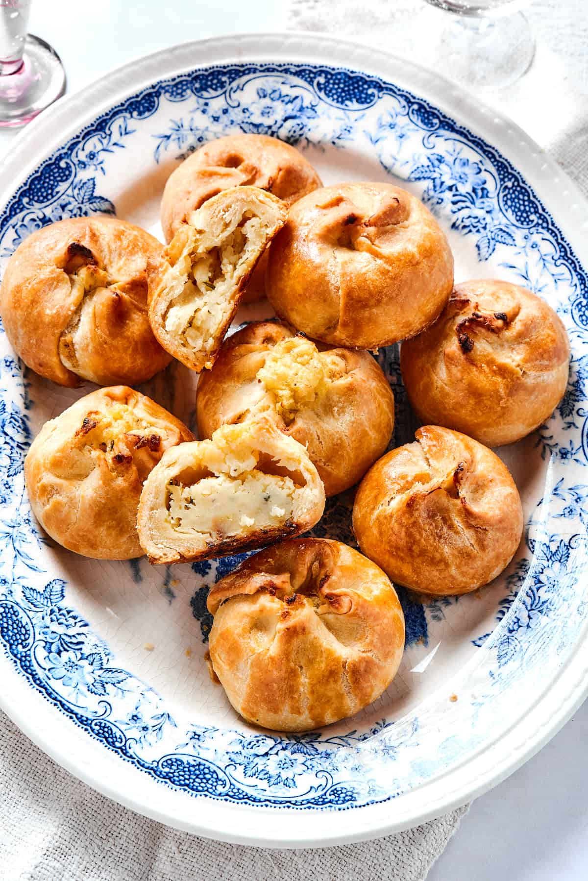 Potato Knishes are a popular Jewish recipe featuring a mashed potato and onion filling wrapped in a thin, flaky dough and baked to golden. Overhead view of a plate of potato knishes, with one cut in half