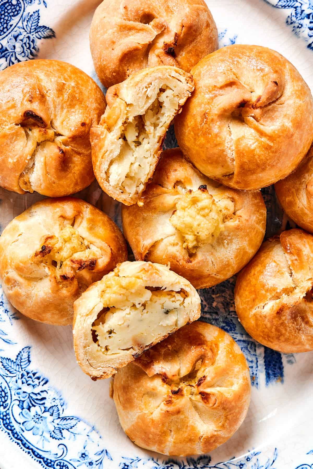 Potato Knishes are a popular Jewish recipe featuring a mashed potato and onion filling wrapped in a thin, flaky dough and baked to golden. Overhead view of a plate of knish, with one cut in half