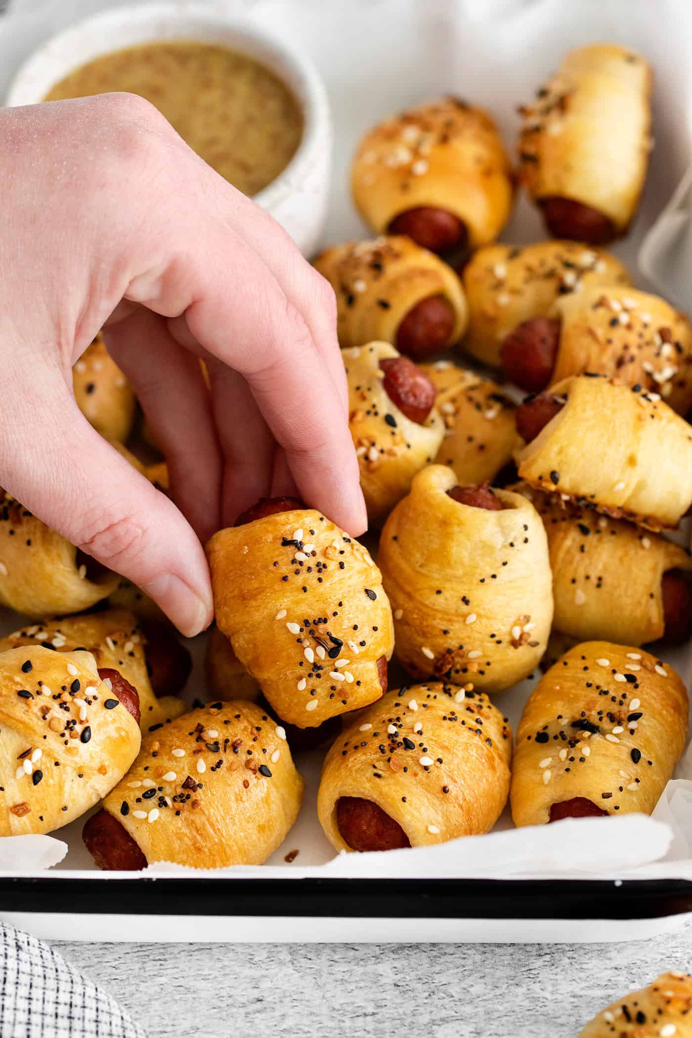 Everything Bagel Pigs in a Blanket are the perfect appetizer– mini sausage wrapped in crescent dough, topped with everything bagel seasoning A hand grabbing a mini pigs in a blanket