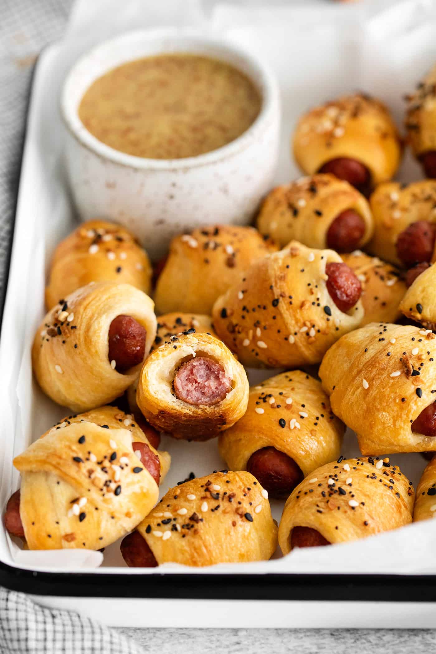 Everything Bagel Pigs in a Blanket are the perfect appetizer– mini sausage wrapped in crescent dough, topped with everything bagel seasoning Mini pigs in a blanket on a tray, one cut in half