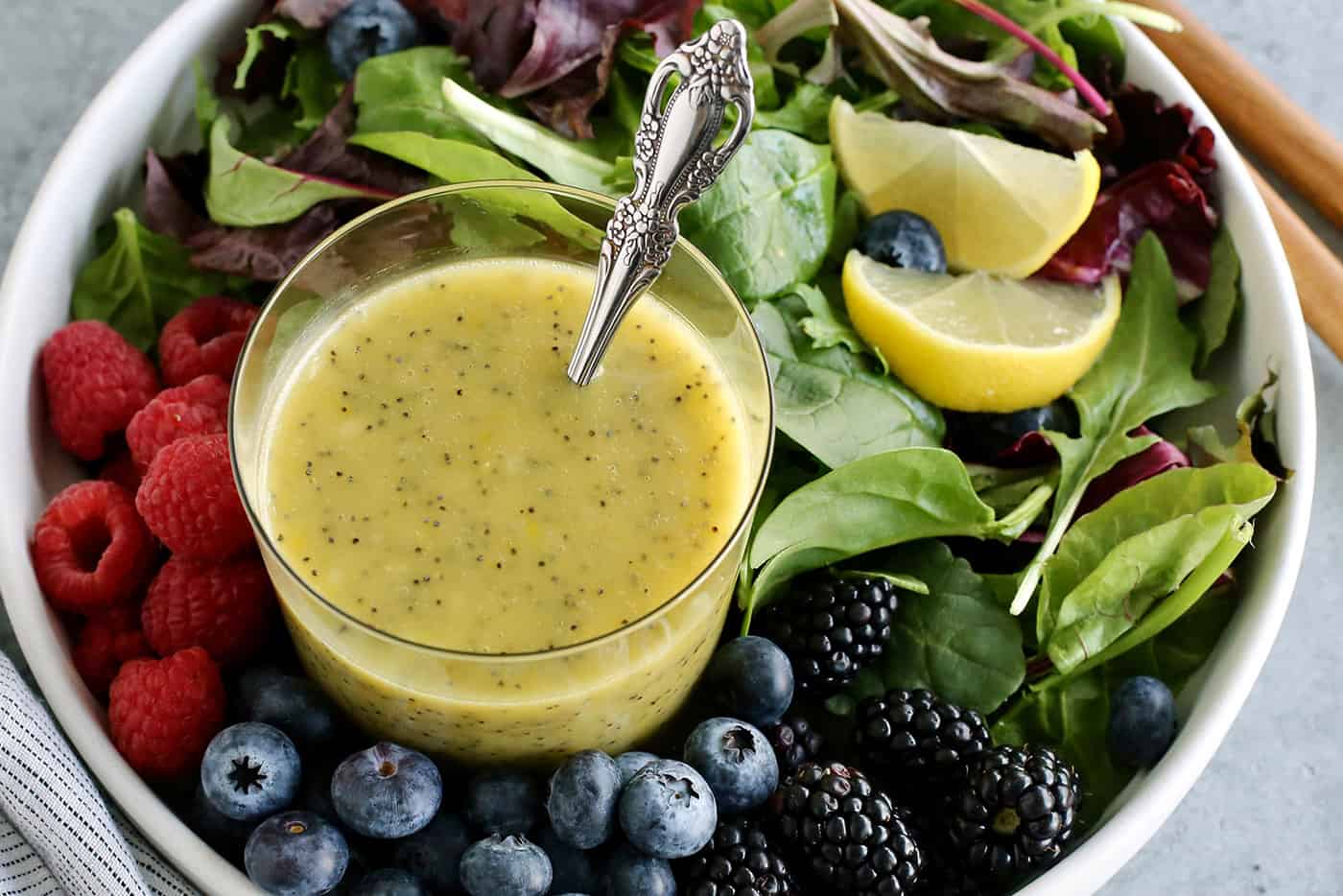 A quick & easy homemade poppy seed dressing with bright lemon flavor and a light, creamy consistency. It's versatile, fresh, and delicious! a cup of poppy seed dressing in a bowl of fresh greens and berries