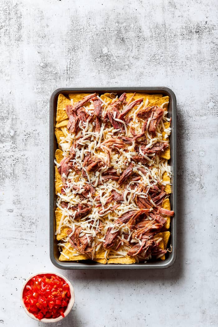 Loaded with pork, cheese, tomatoes, and all the toppings you desire, these Pulled Pork Nachos are an easy and tasty appetizer. Tortilla chips topped with pulled pork on a baking sheet