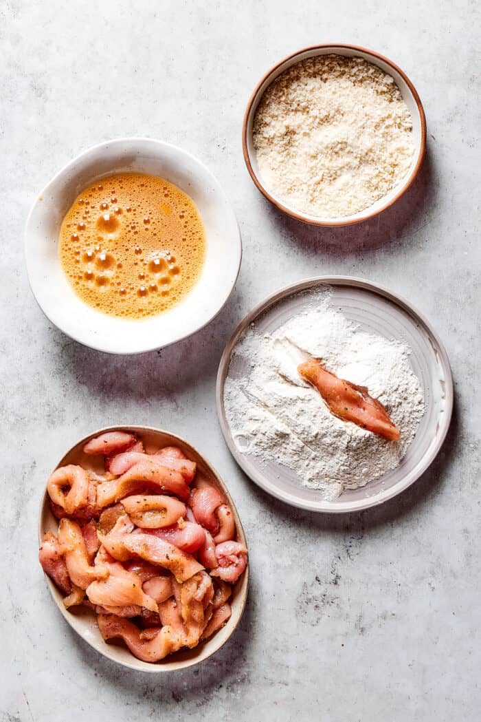 Made with thin, perfectly seasoned strips of chicken, these Chicken Fries are perfect dipped in this tangy roasted garlic aioli. A plate of seasoned chicken strips, a bowl of flour, a bowl of breadcrumbs, and egg wash