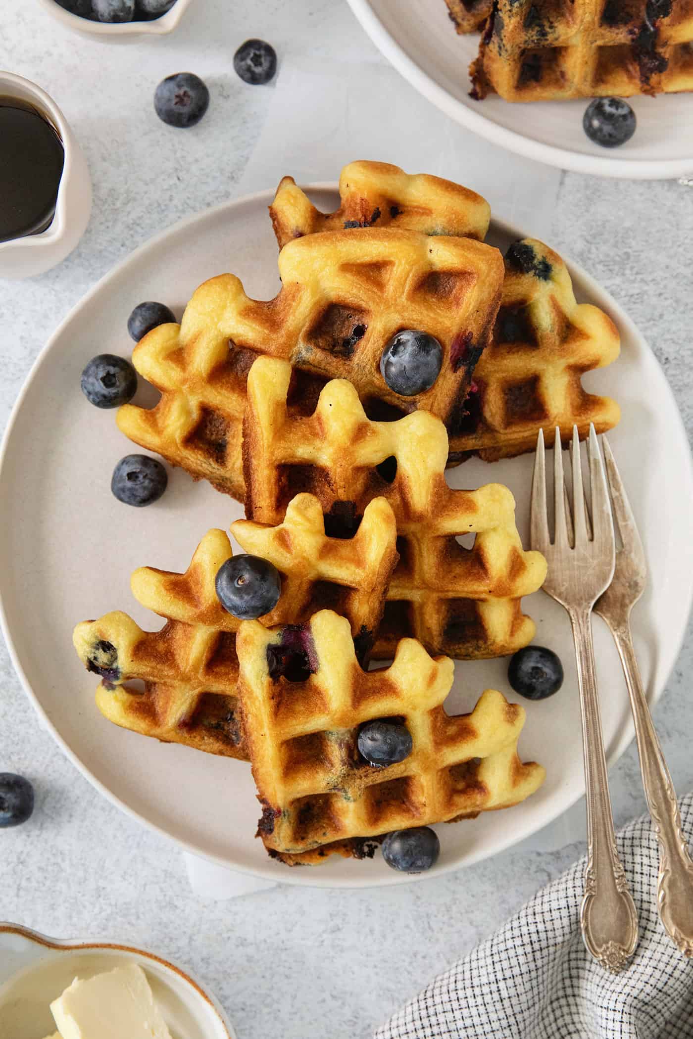 Simple and wholesome, these Blueberry Waffles are sure to please. Serve them to Mom on her special day or make them for the whole family! Blueberry waffles topped with blueberries are shown on a white plate with forks.