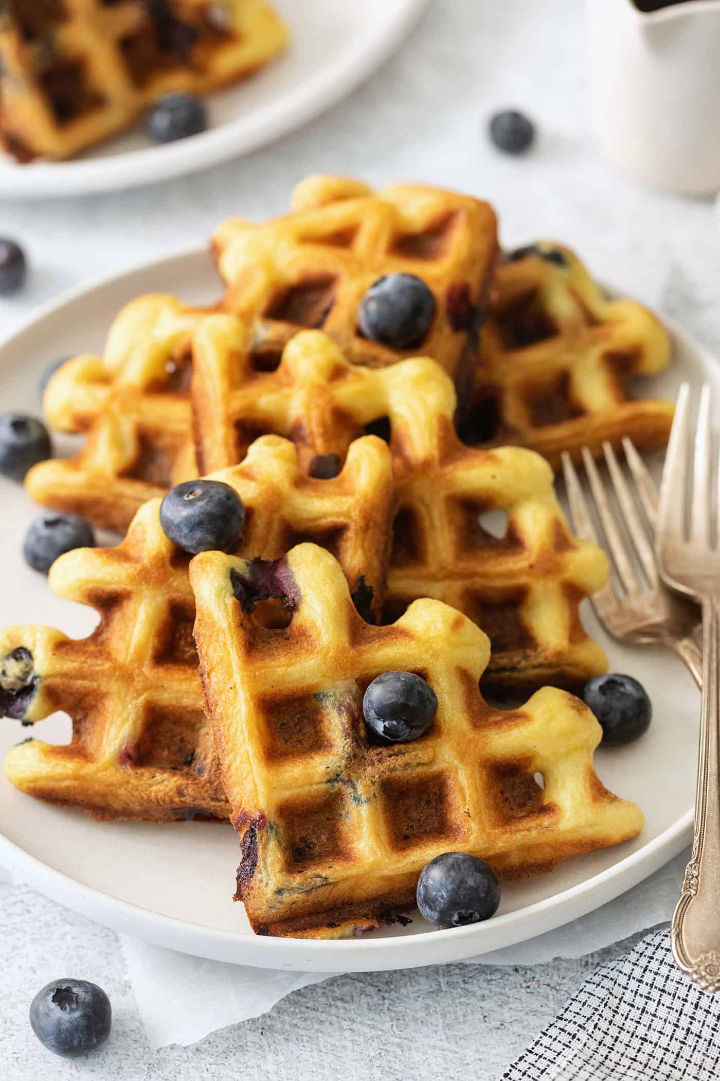 Simple and wholesome, these Blueberry Waffles are sure to please. Serve them to Mom on her special day or make them for the whole family! Blueberry waffles topped with blueberries are shown on a white plate with forks.