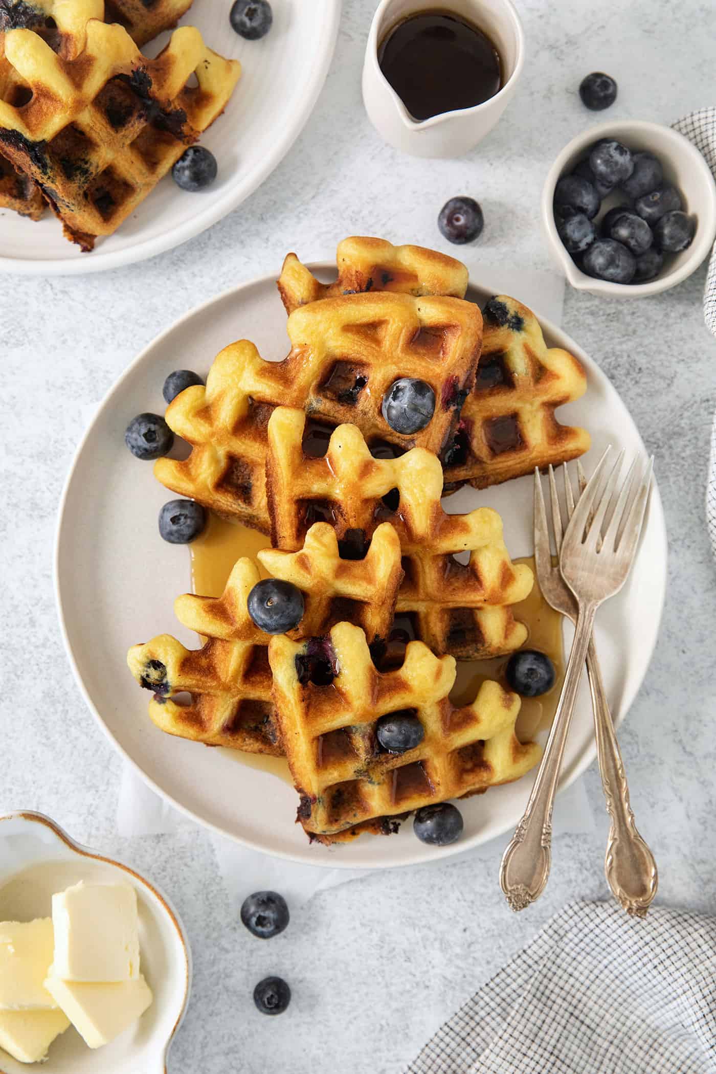 Simple and wholesome, these Blueberry Waffles are sure to please. Serve them to Mom on her special day or make them for the whole family! Blueberry waffles topped with blueberries are shown on a white plate with forks.