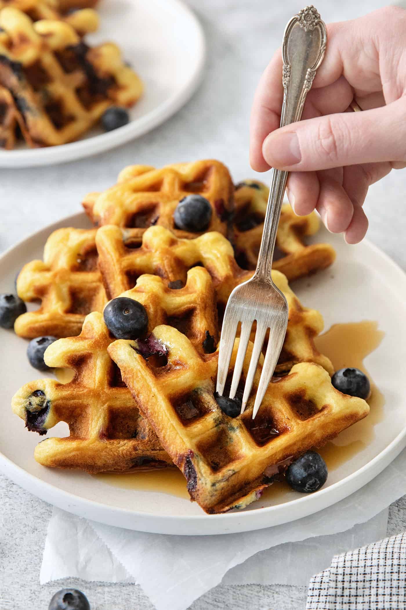 Simple and wholesome, these Blueberry Waffles are sure to please. Serve them to Mom on her special day or make them for the whole family! A hand holds a fork to cup into a plate of blueberry waffles.