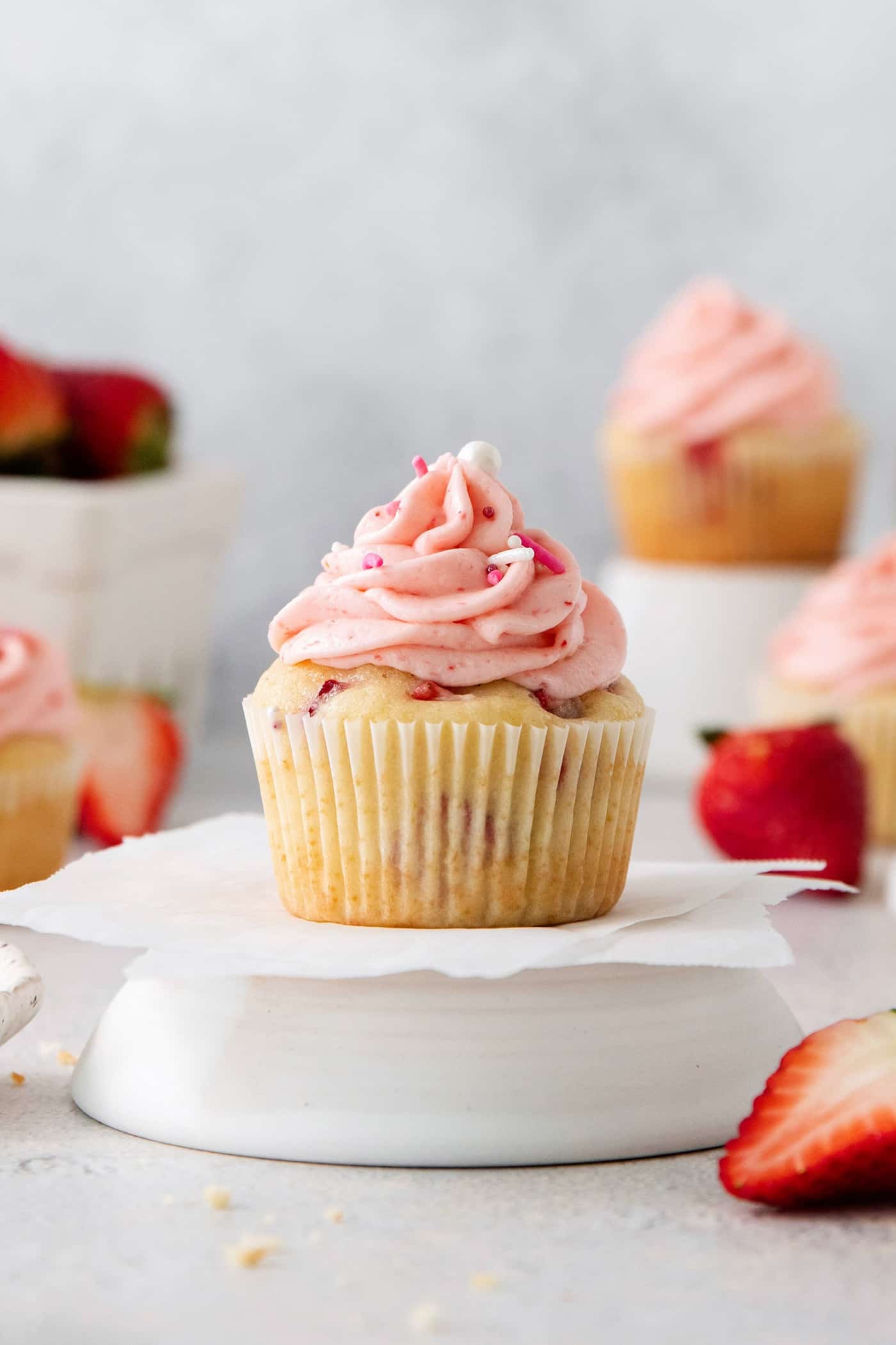 Vibrantly pink Strawberry Cupcakes will dress up your Mother's Day table - or make them anytime for a special strawberry treat. A frosted strawberry cupcake is show on a cupcake stand with cupcakes and strawberries in the background.
