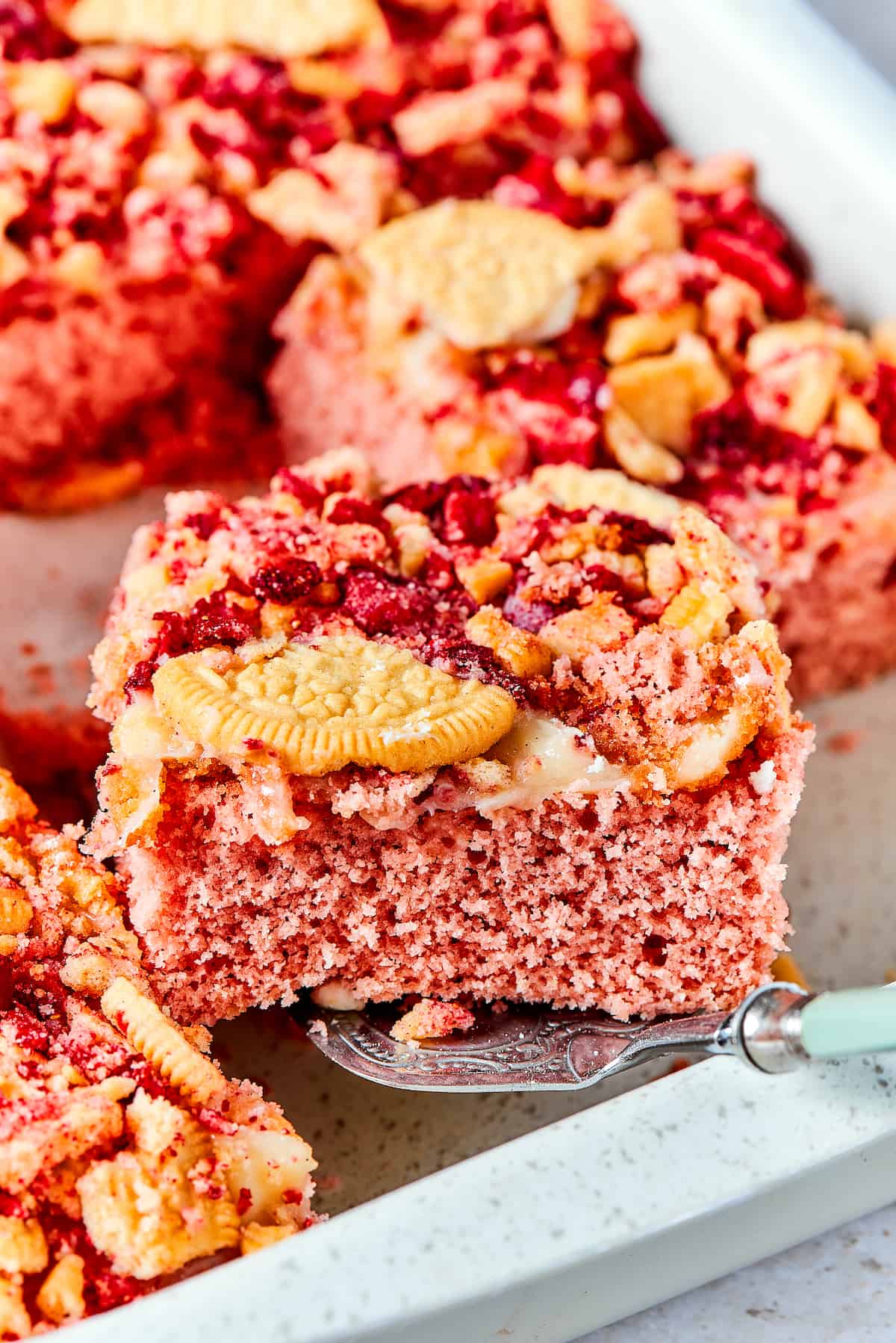 Creamy and sweet with a bit of crunch, Strawberry Crunch Cake is bursting with fruit flavor! This bright strawberry dessert is a favorite. A piece of strawberry crunch cake is lifted out of a pan.