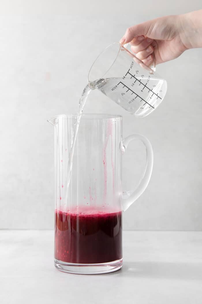 This Blueberry Lemonade is the perfect summertime drink! It’s fruity and refreshing with tart lemons and sweet blueberries. Water being added to a pitcher with lemon juice and blueberry syrup