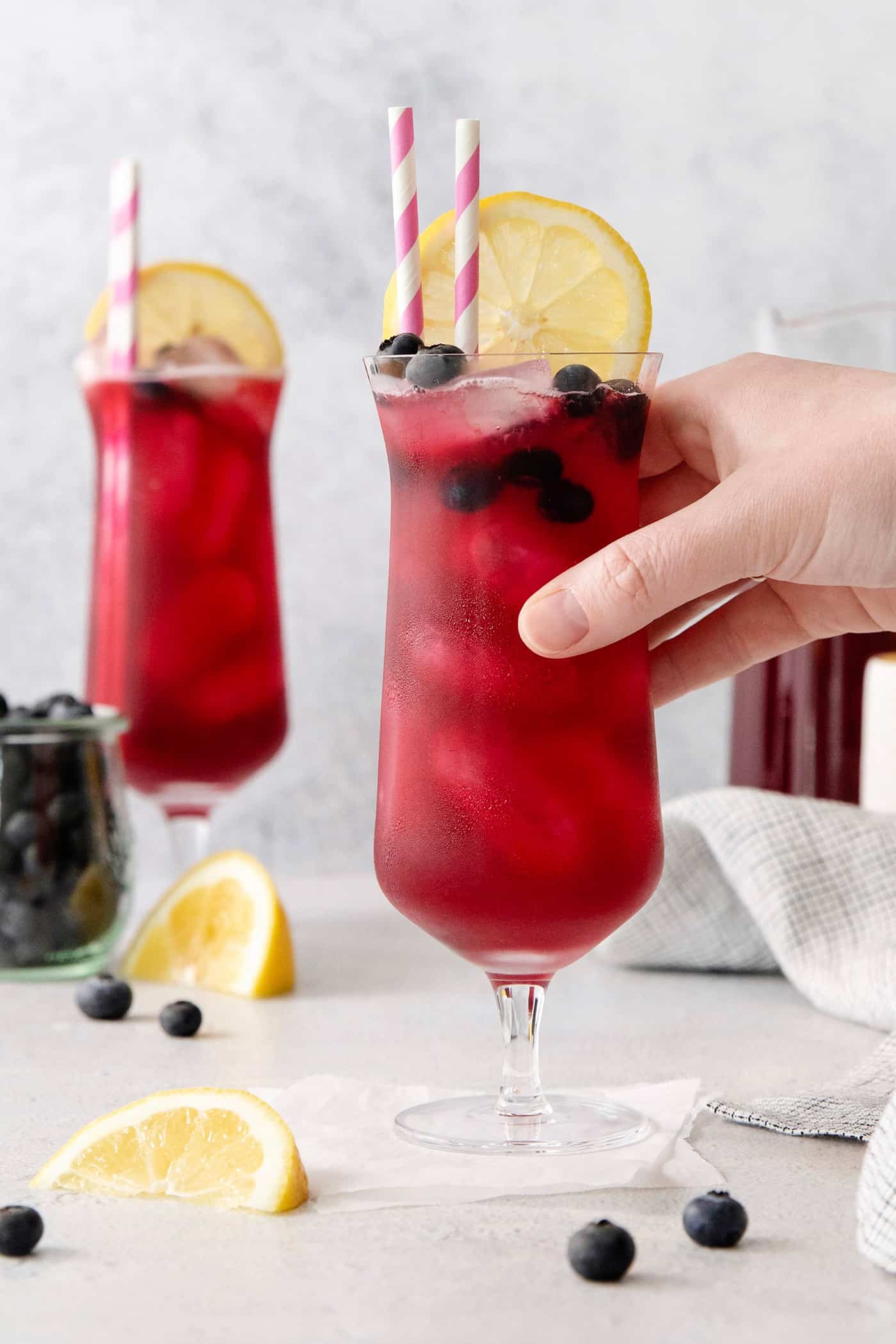 This Blueberry Lemonade is the perfect summertime drink! It’s fruity and refreshing with tart lemons and sweet blueberries. A hand grabbing a glass of blueberry lemonade
