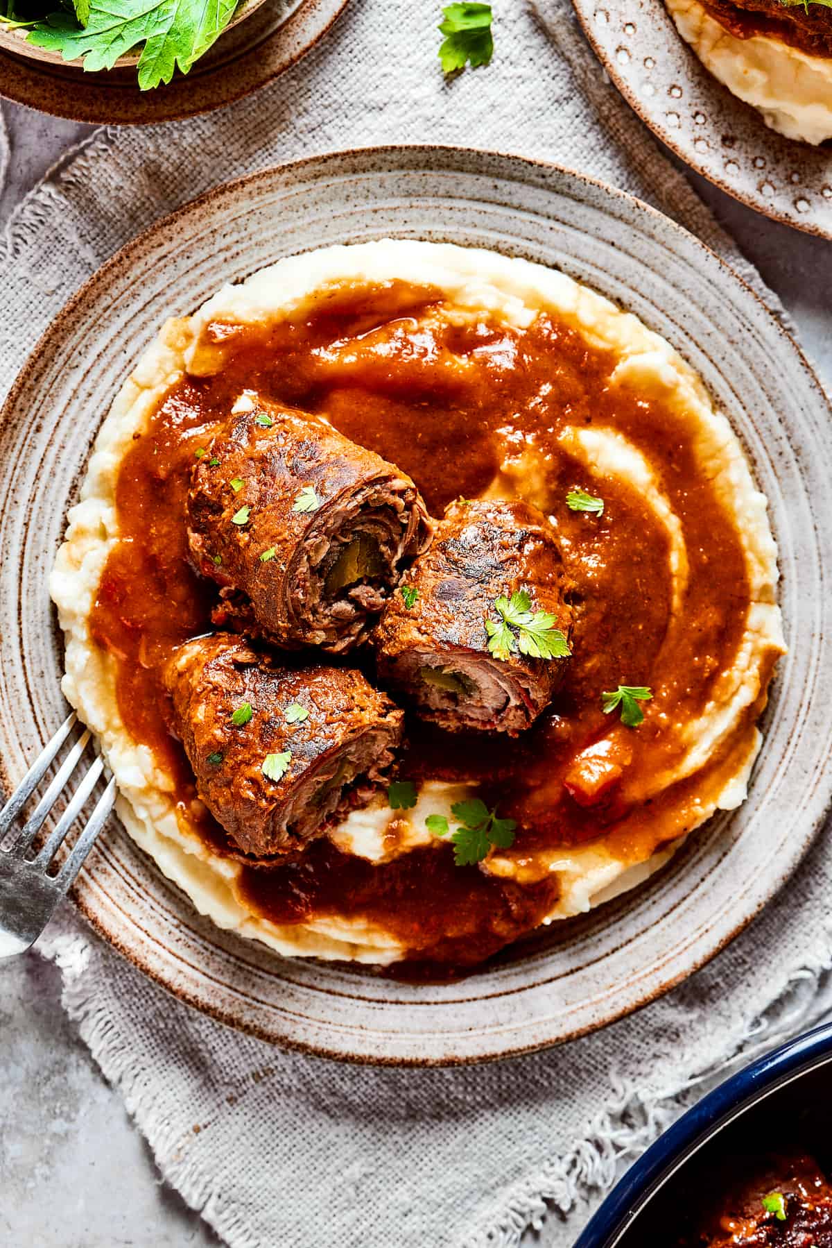 Rich, flavorful Rouladen is a classic German dish. Little meat rolls are stuffed with onions, bacon, and pickles then simmered in gravy. Rouladen on top of mashed potatoes on a plate.