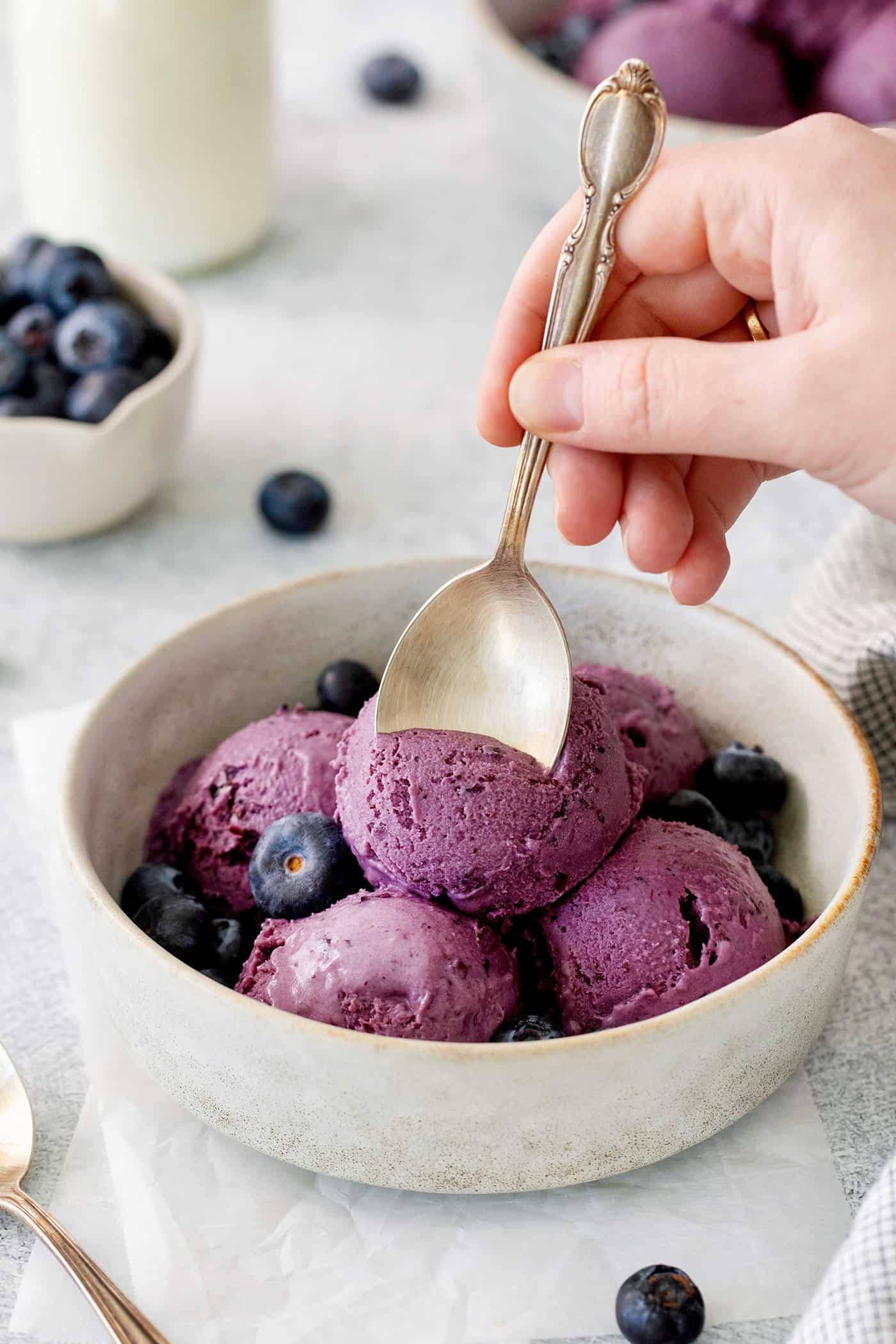 Summer-sweet Crème Fraîche Blueberry Ice Cream swirls roasted blueberries into a creamy ice cream to make the best of blueberry season! Blueberry ice cream in a bowl with a hand and a spoon.
