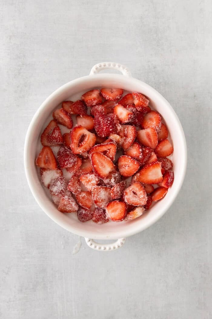 This homemade Crème Fraîche Strawberry Ice Cream is rich and ultra creamy with a gorgeous strawberry swirl folded into a tangy ice cream. Strawberries mixed with sugar in a white bowl.