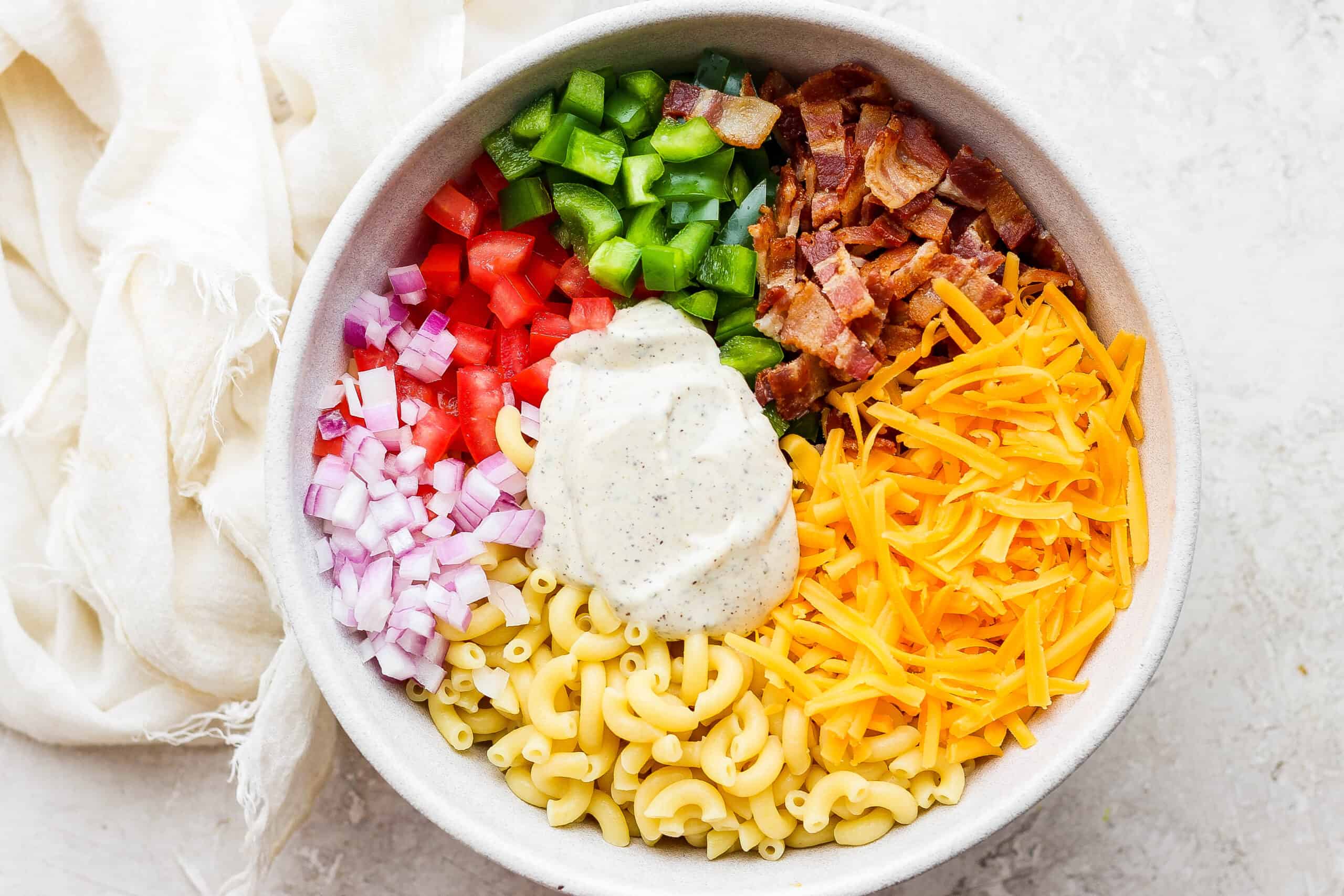 Make this crowd-pleasing Bacon Ranch Pasta Salad for your next bbq! Crispy bacon, and tender pasta will make this potluck side a new favorite. Dressing is shown on top of a white bowl of bacon ranch pasta salad.