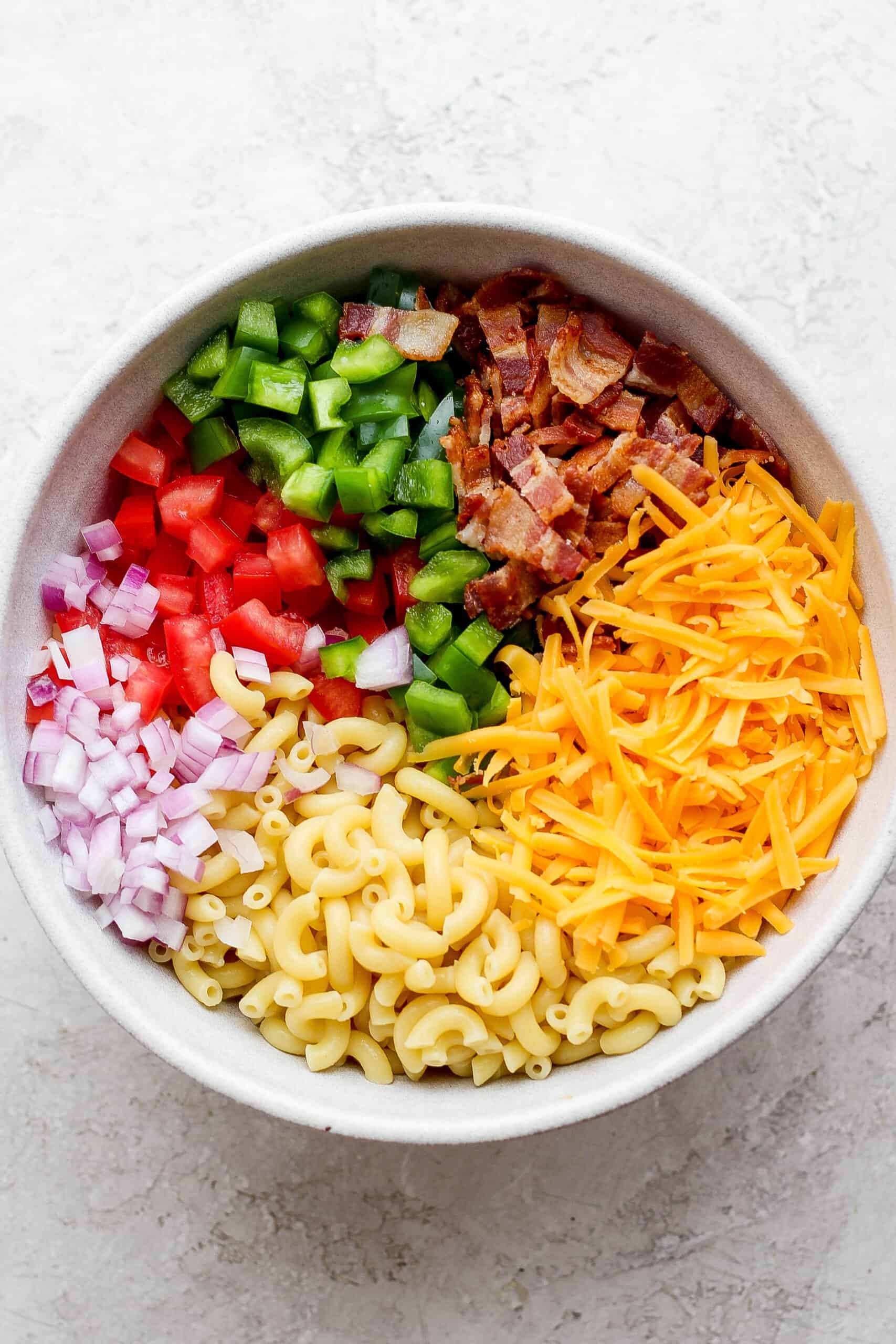 Make this crowd-pleasing Bacon Ranch Pasta Salad for your next bbq! Crispy bacon, and tender pasta will make this potluck side a new favorite. Components of a bacon ranch pasta salad including green onions, red onions, macaroni, cheddar cheese, pepper, and bacon are shown in a white bowl.