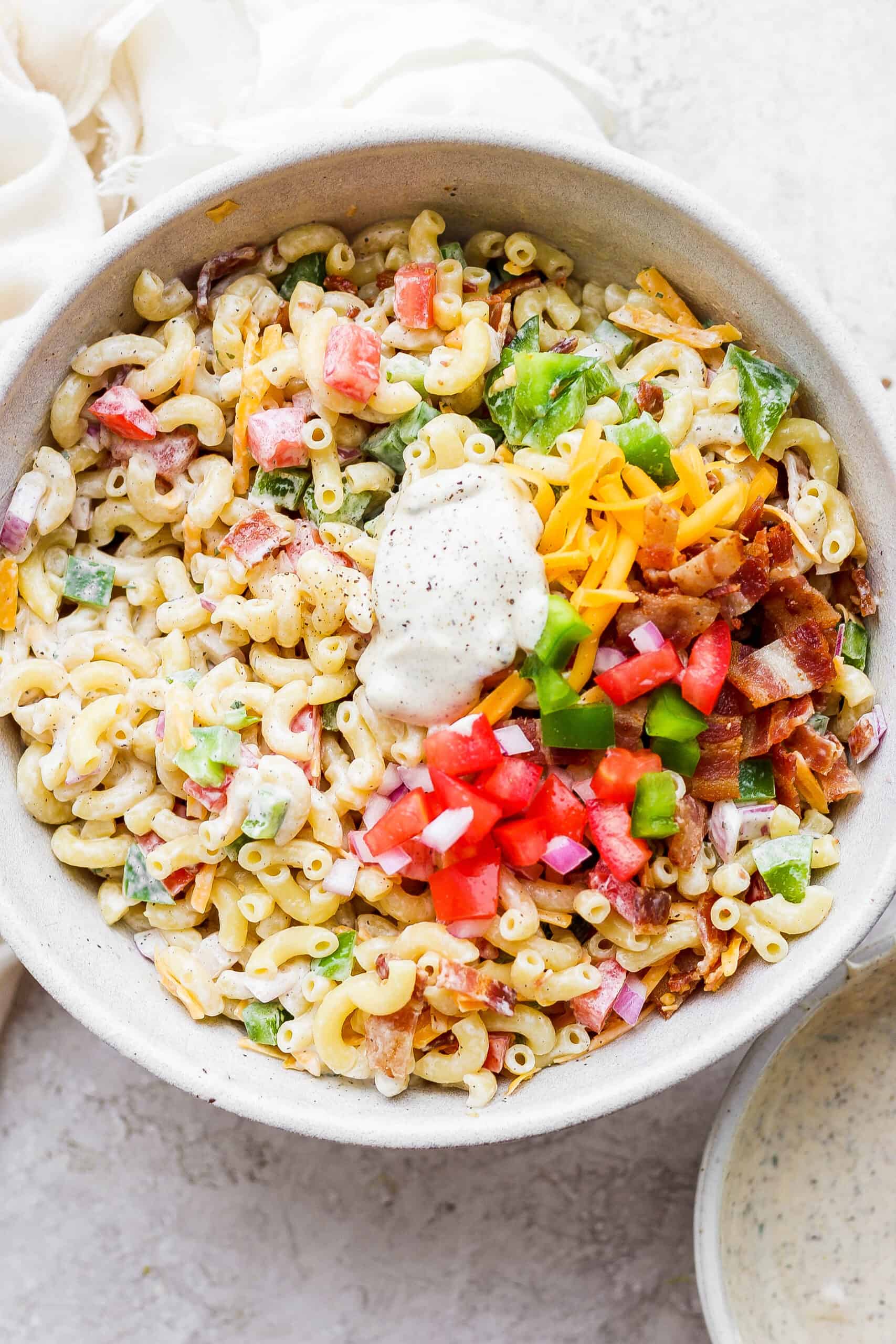 Make this crowd-pleasing Bacon Ranch Pasta Salad for your next bbq! Crispy bacon, and tender pasta will make this potluck side a new favorite. Vegetables, bacon, and macaroni shown in a white bowl with dressing on top.