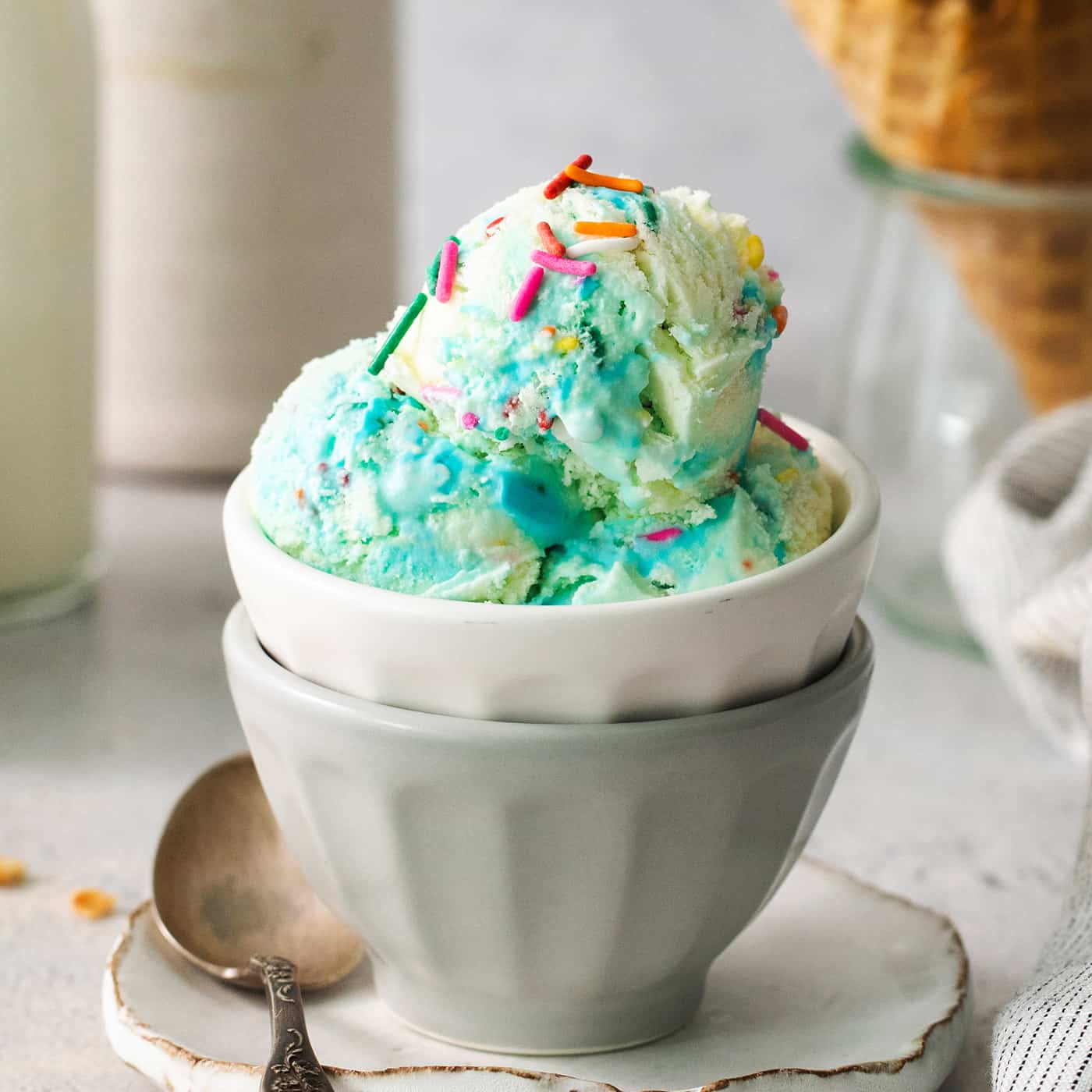 This Birthday Cake Ice Cream is delightfully rich and creamy, with swirls of blue vanilla frosting and sprinkles. No custard required! A bowl of blue birthday cake ice cream with a cone of ice cream next to it.