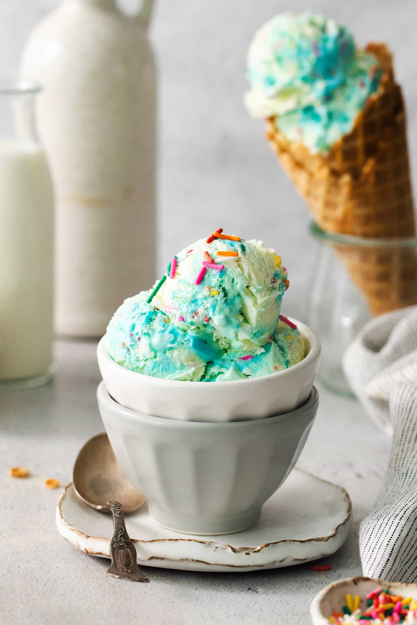 This Birthday Cake Ice Cream is delightfully rich and creamy, with swirls of blue vanilla frosting and sprinkles. No custard required! A bowl of blue birthday cake ice cream with a cone of ice cream next to it.