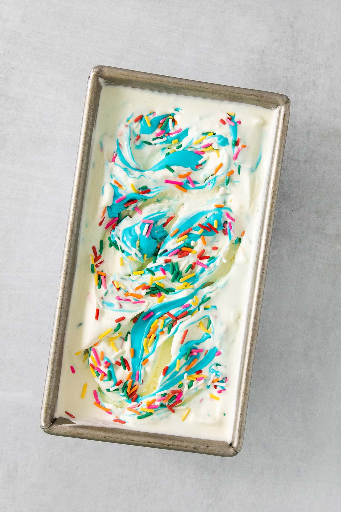This Birthday Cake Ice Cream is delightfully rich and creamy, with swirls of blue vanilla frosting and sprinkles. No custard required! A loaf pan with birthday cake ice cream topped with blue frosting and sprinkles.