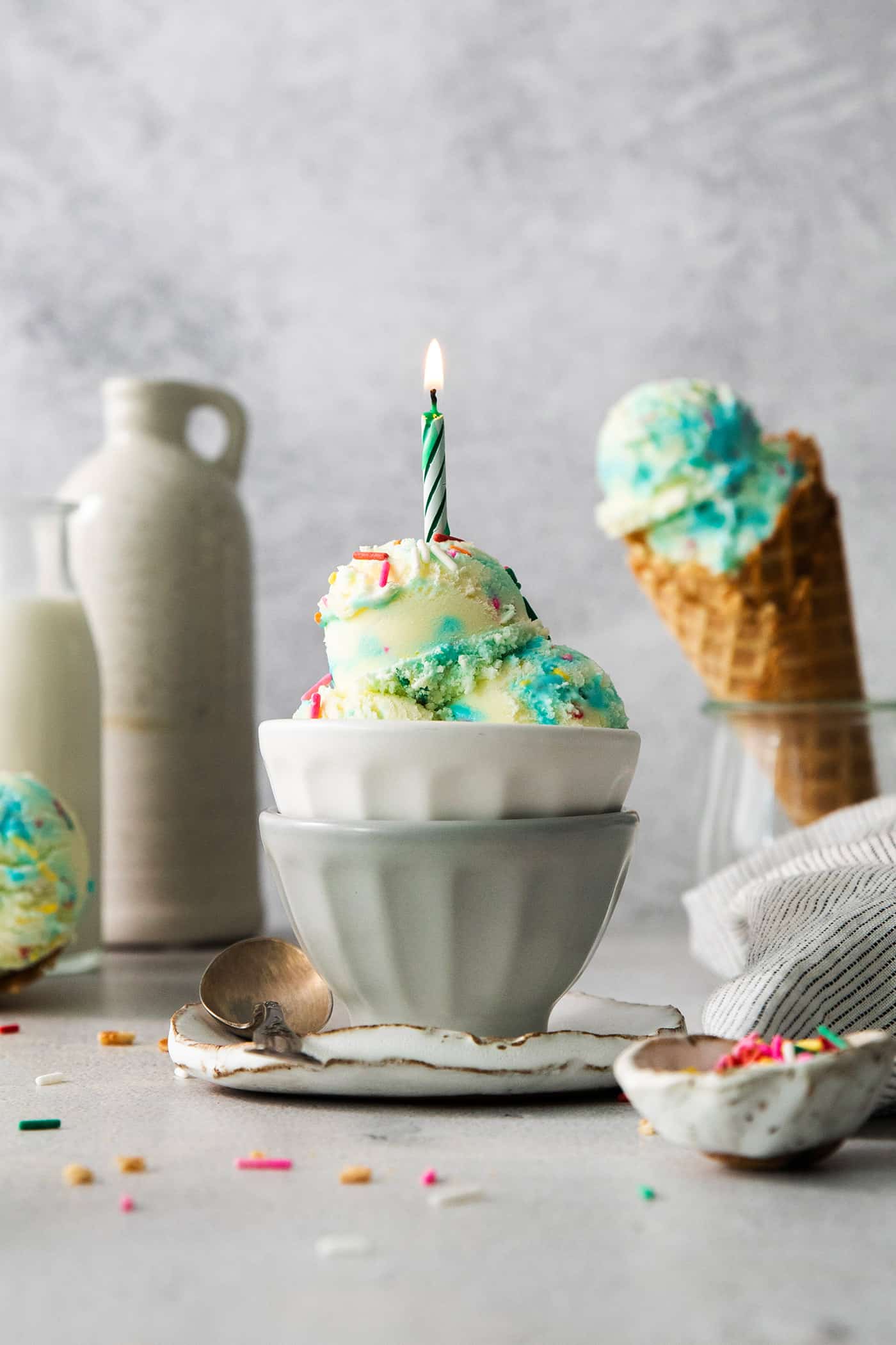 This Birthday Cake Ice Cream is delightfully rich and creamy, with swirls of blue vanilla frosting and sprinkles. No custard required! A bowl of birthday cake ice cream topped with a burning candle with a cone of ice cream next to it.