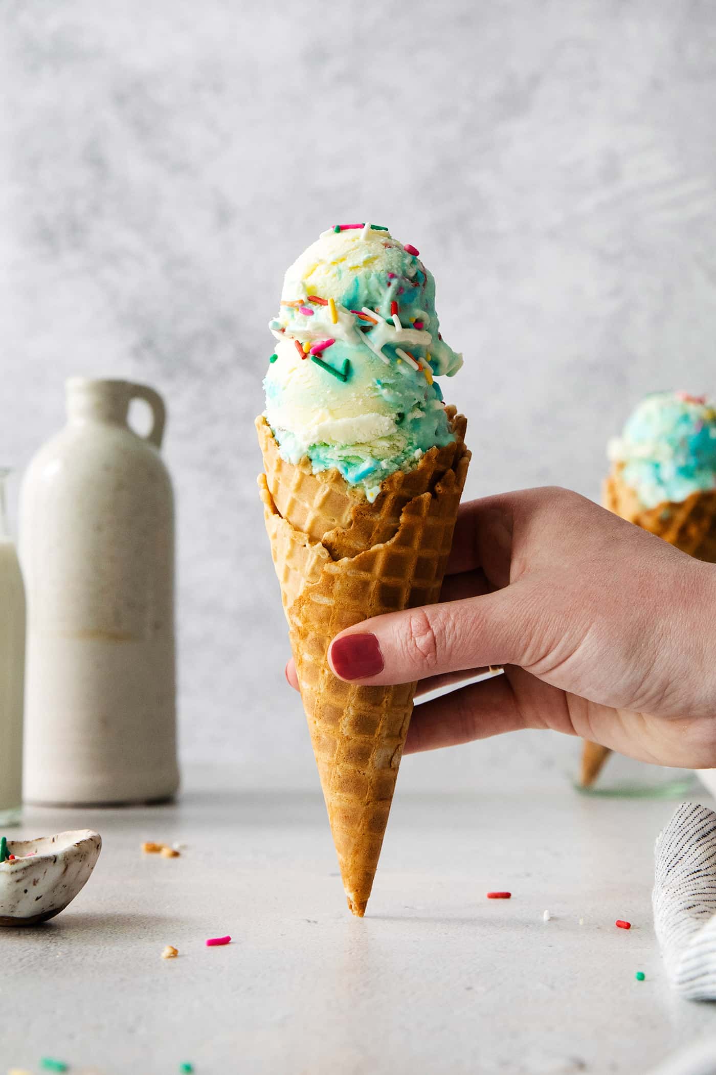 This Birthday Cake Ice Cream is delightfully rich and creamy, with swirls of blue vanilla frosting and sprinkles. No custard required! A hand holds a cone of birthday cake ice cream.