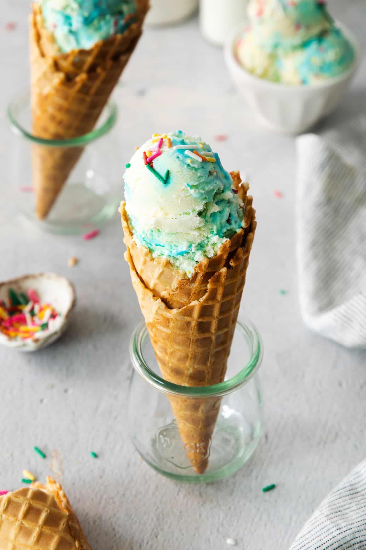 This Birthday Cake Ice Cream is delightfully rich and creamy, with swirls of blue vanilla frosting and sprinkles. No custard required! Birthday cake ice cream cones with rainbow sprinkles alongside.