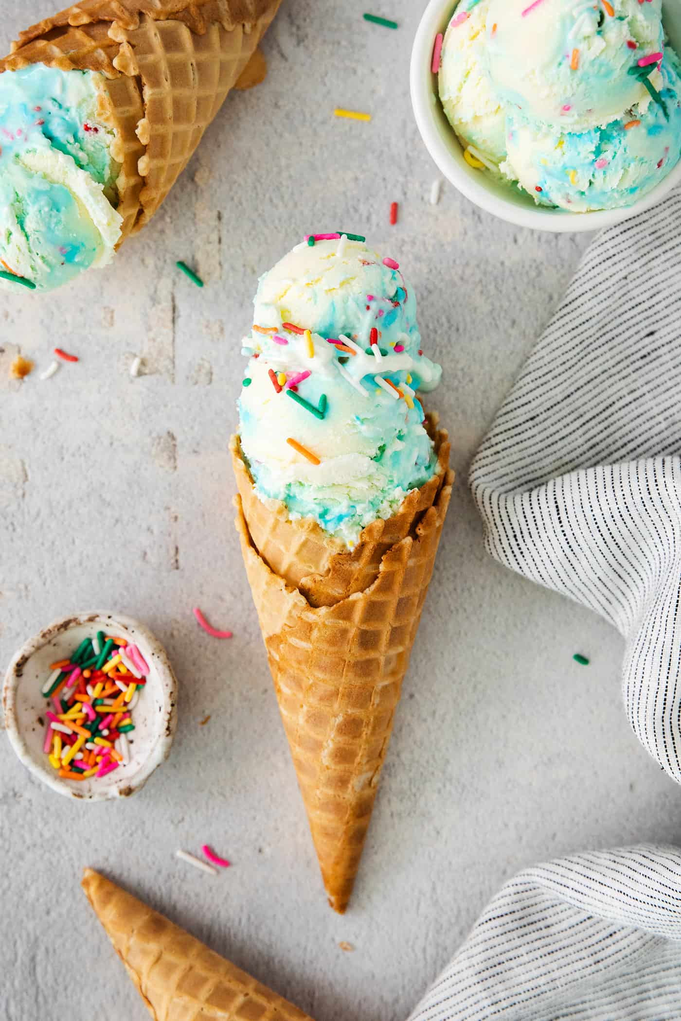 This Birthday Cake Ice Cream is delightfully rich and creamy, with swirls of blue vanilla frosting and sprinkles. No custard required! Birthday cake ice cream cones with rainbow sprinkles alongside.
