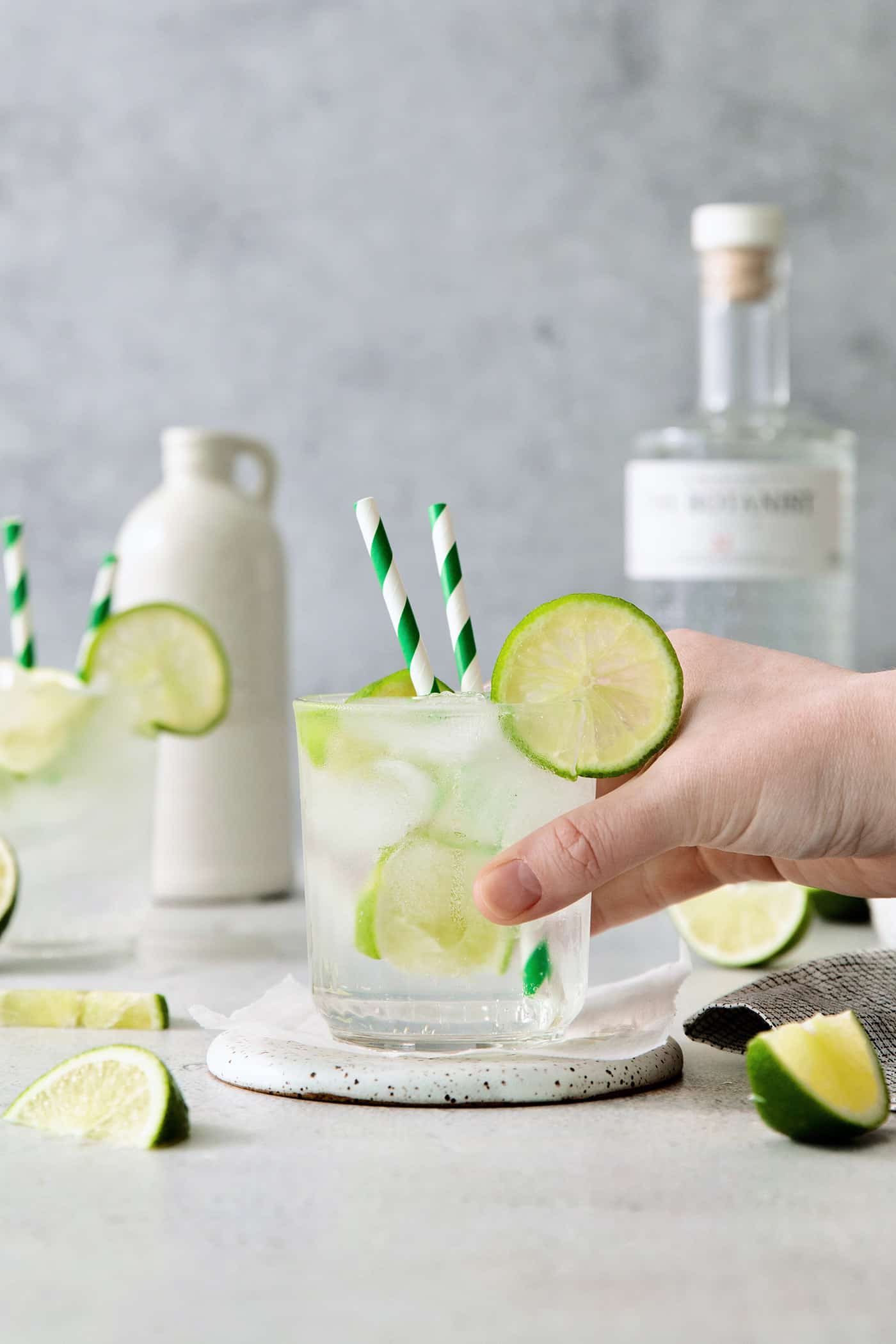 A Classic Gin and Tonic is a must to master. This recipe is for a super simple, super refreshing, and always-in-style cocktail. A hand picks up a glass of gin and tonic with two straws and lime circles.