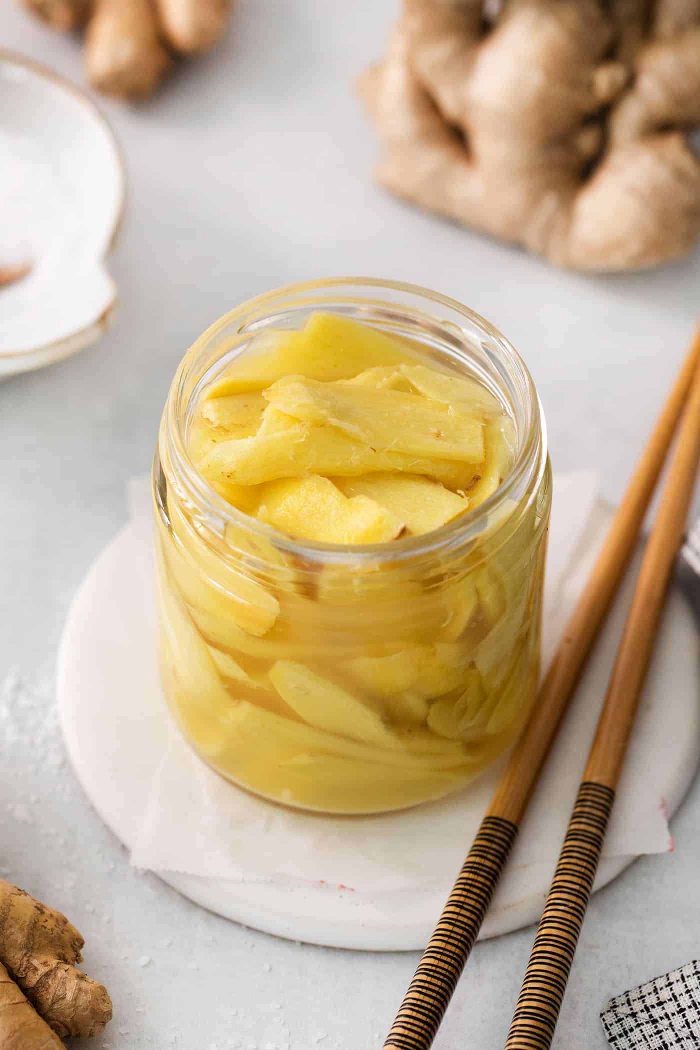 Peppery, pink Pickled Ginger is a wonderful companion to sushi and it's so easy to make at home! You need just 5 ingredients. A jar of pickled ginger on a white plate with chopsticks next to it.